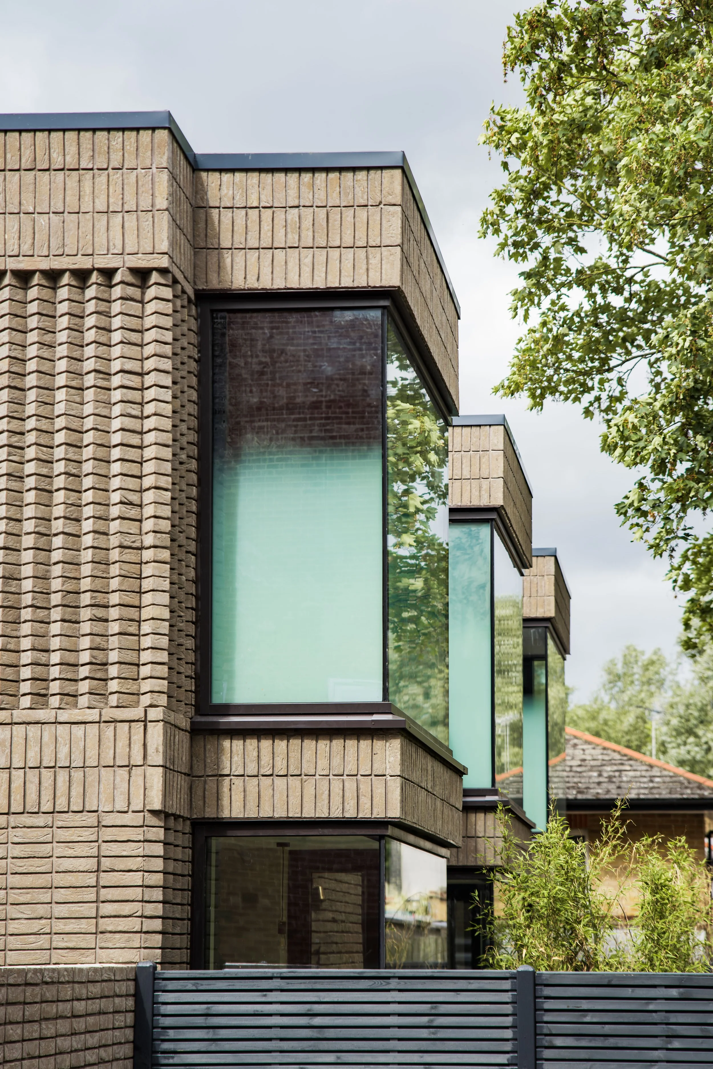 Close-up view of a modern building with beige brick walls and large glass windows, accompanied by green trees and shrubs in the surroundings.