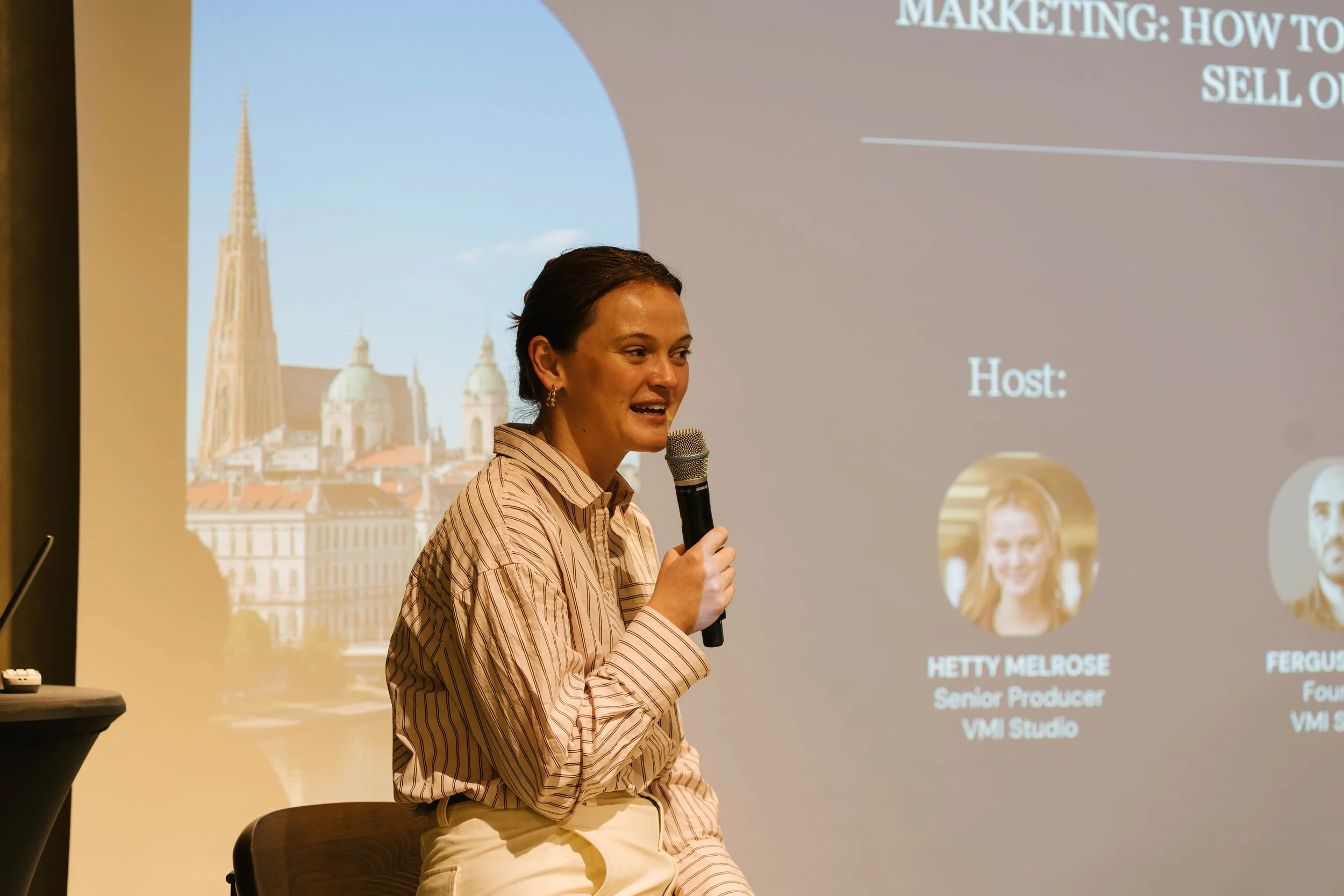 A woman with dark hair tied back, wearing a beige striped shirt, is speaking into a handheld microphone while sitting on a chair. Behind her, a presentation slide is displayed with text about marketing and sales, and there is a background image of a 