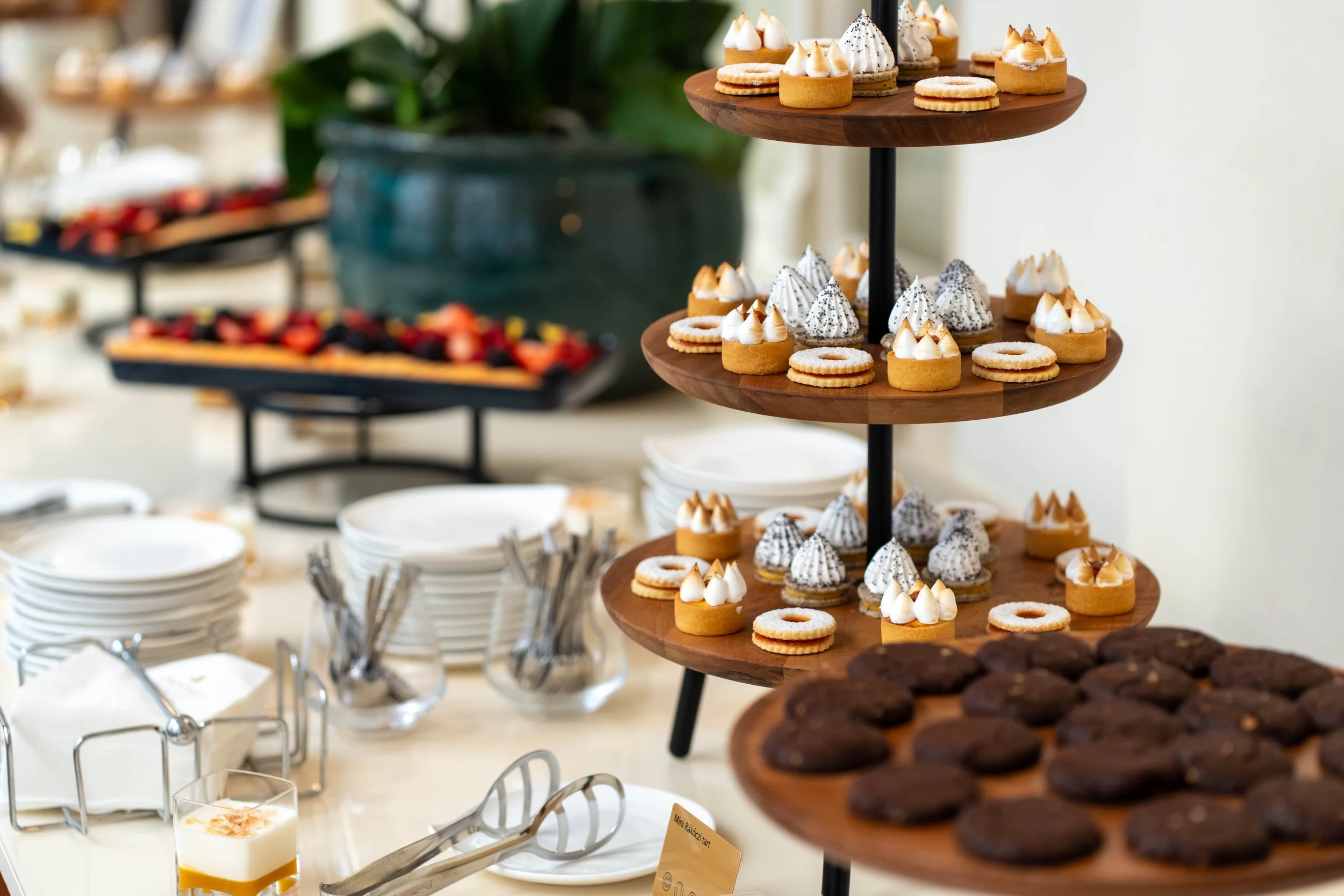 Tiered wooden stand with assorted small desserts including tarts topped with whipped cream, cookies, and chocolates, at a dessert buffet with plates and glasses in the background.