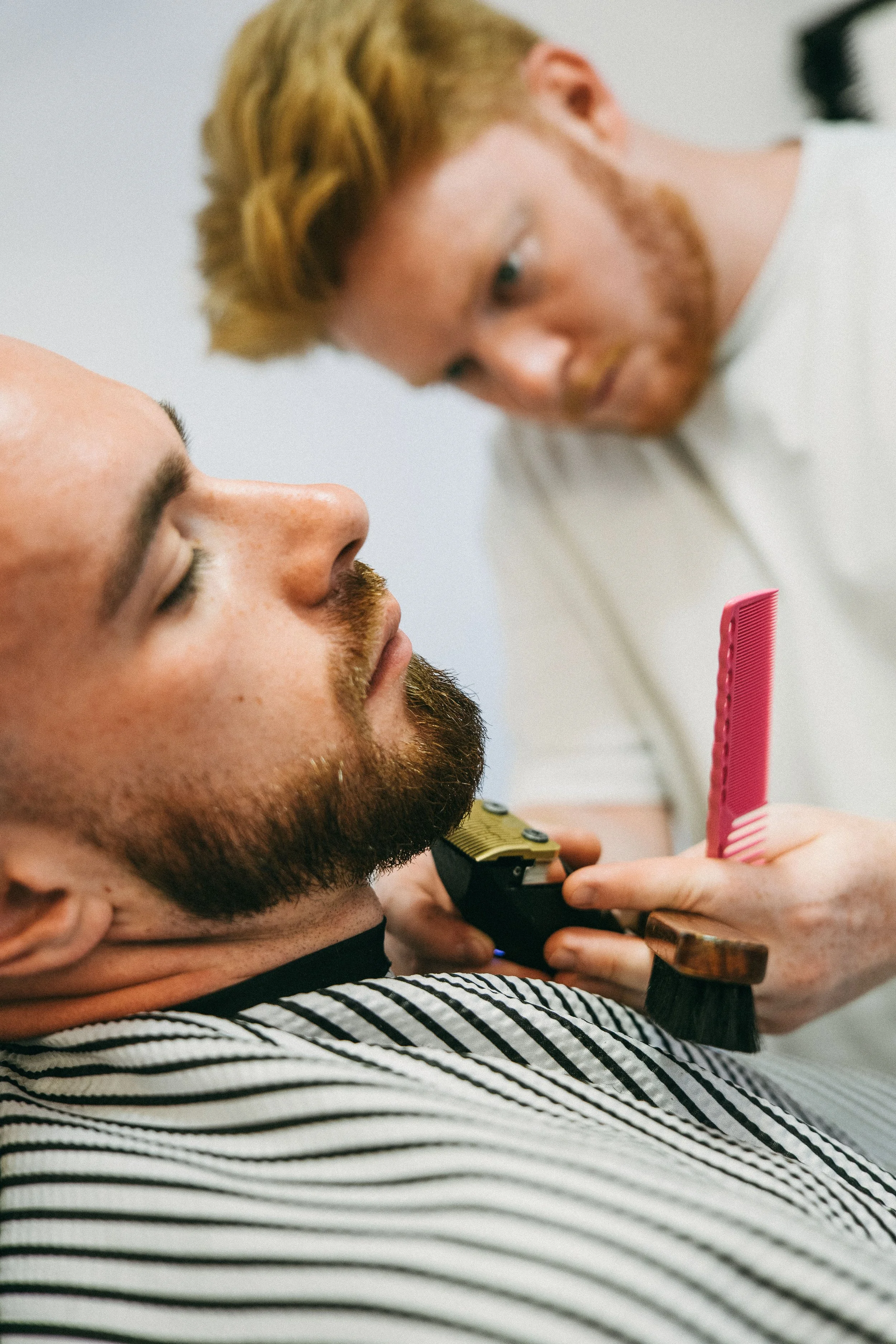 A man with a beard and a striped shirt is getting a haircut, while a barber with blonde hair and glasses is using scissors and a comb to trim his hair.