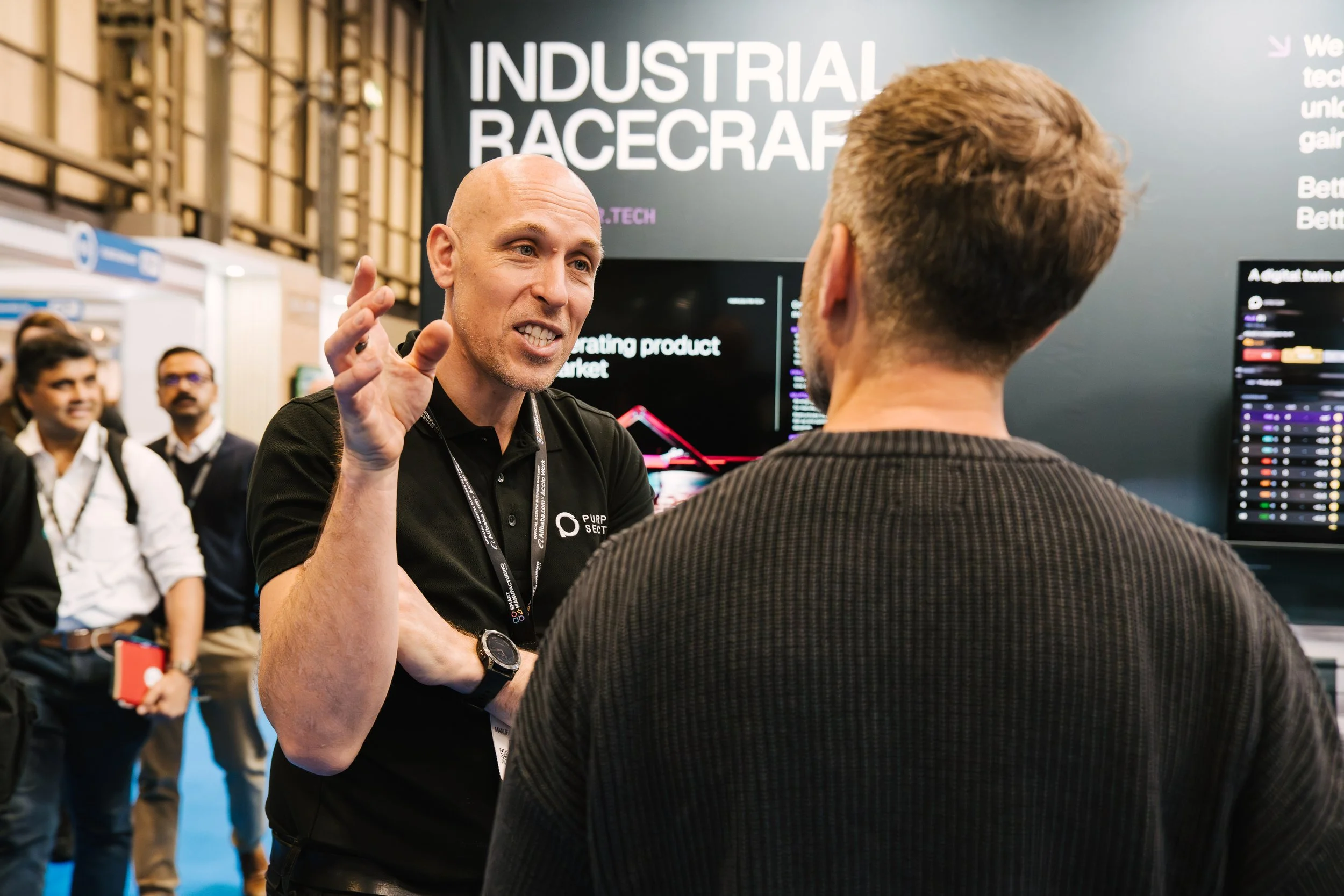 At an industrial racecraft event, a bald man in a black shirt is speaking animatedly to another man with curly hair. In the background, there are event attendees, a large black sign with white text reading "INDUSTRIAL RACECRAFT," and electronic displ