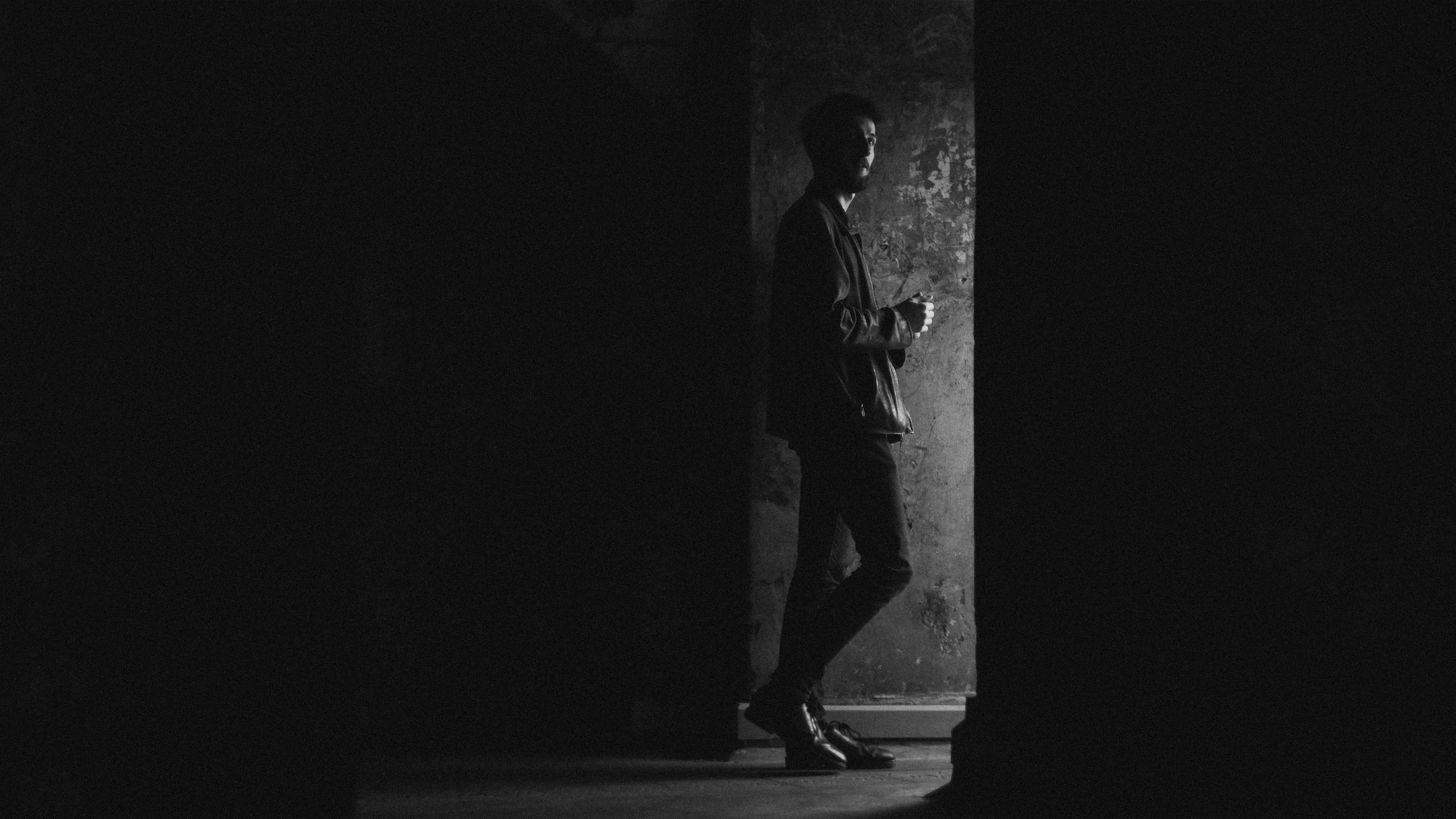 A person standing near a doorway, partially hidden in shadow, wearing dark clothing and high-heeled boots, holding a cup, with textured wall behind.