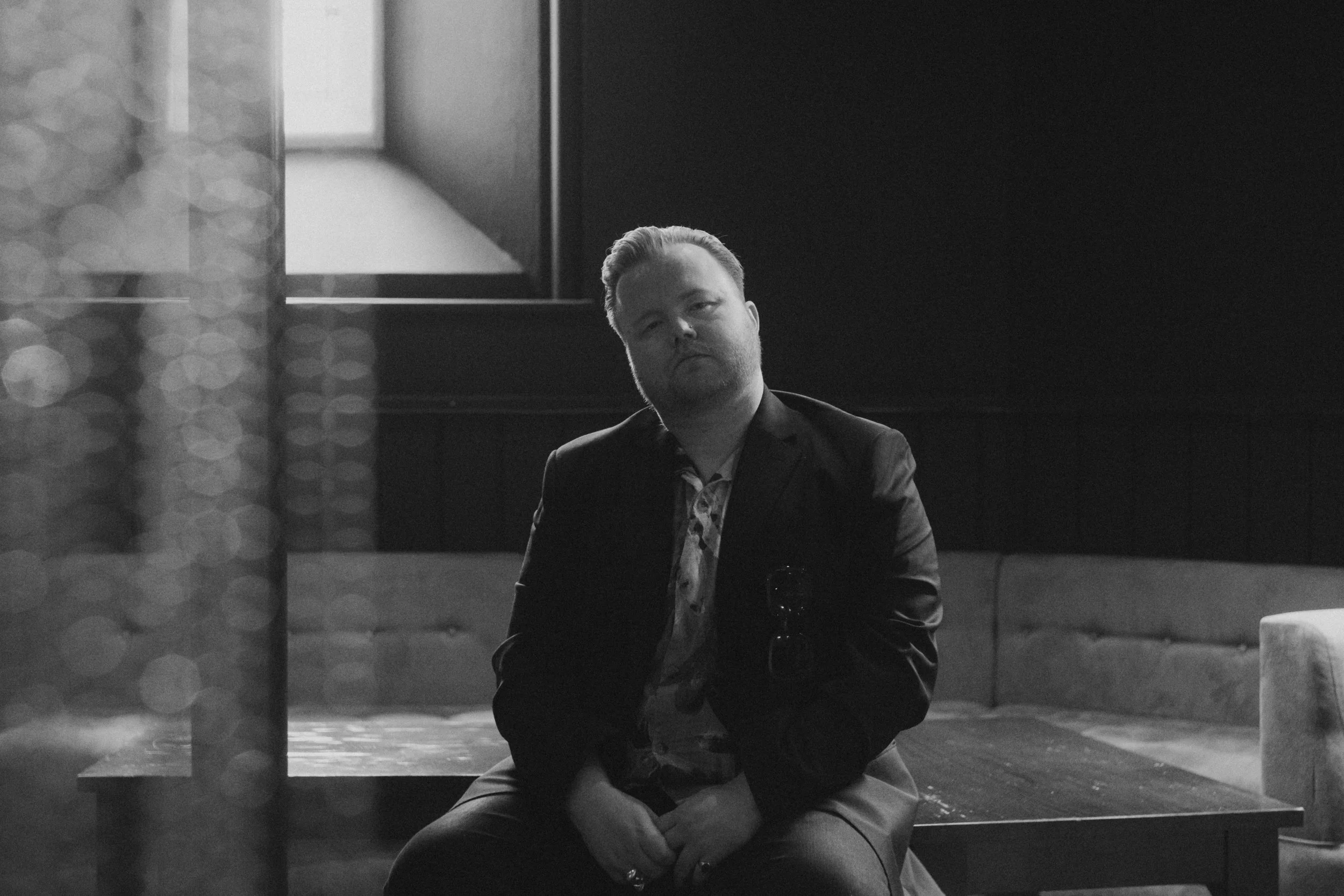 A black-and-white photo of a man sitting outside, dressed in a blazer and patterned shirt, looking directly at the camera with a neutral expression. The background features a brick wall and a window, with soft lighting and bokeh effects in the foregr