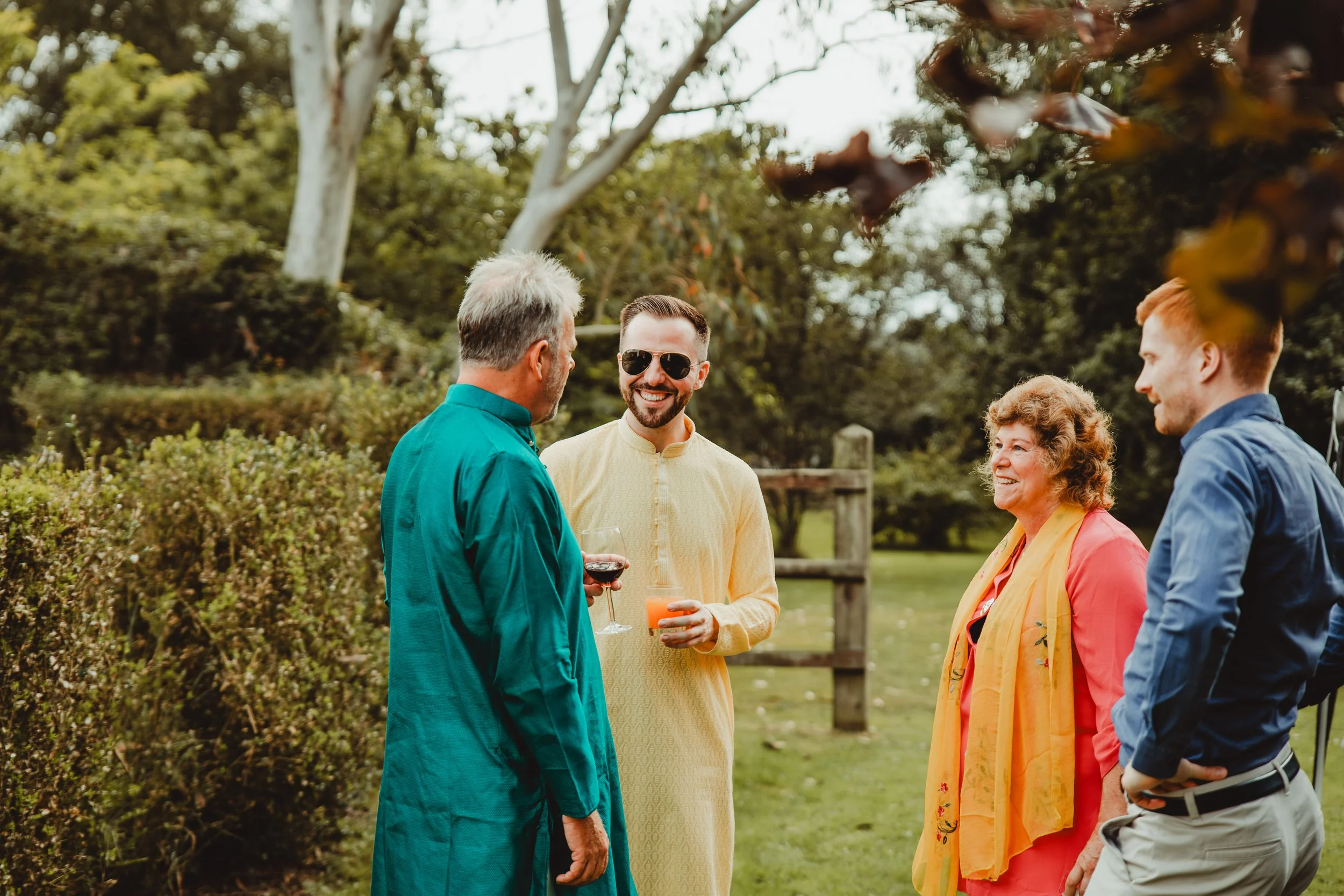A group of five adults outdoors, engaging in conversation. One person is holding a glass of red wine, another a drink with orange juice. They are surrounded by trees and greenery, suggesting a garden or park setting.
