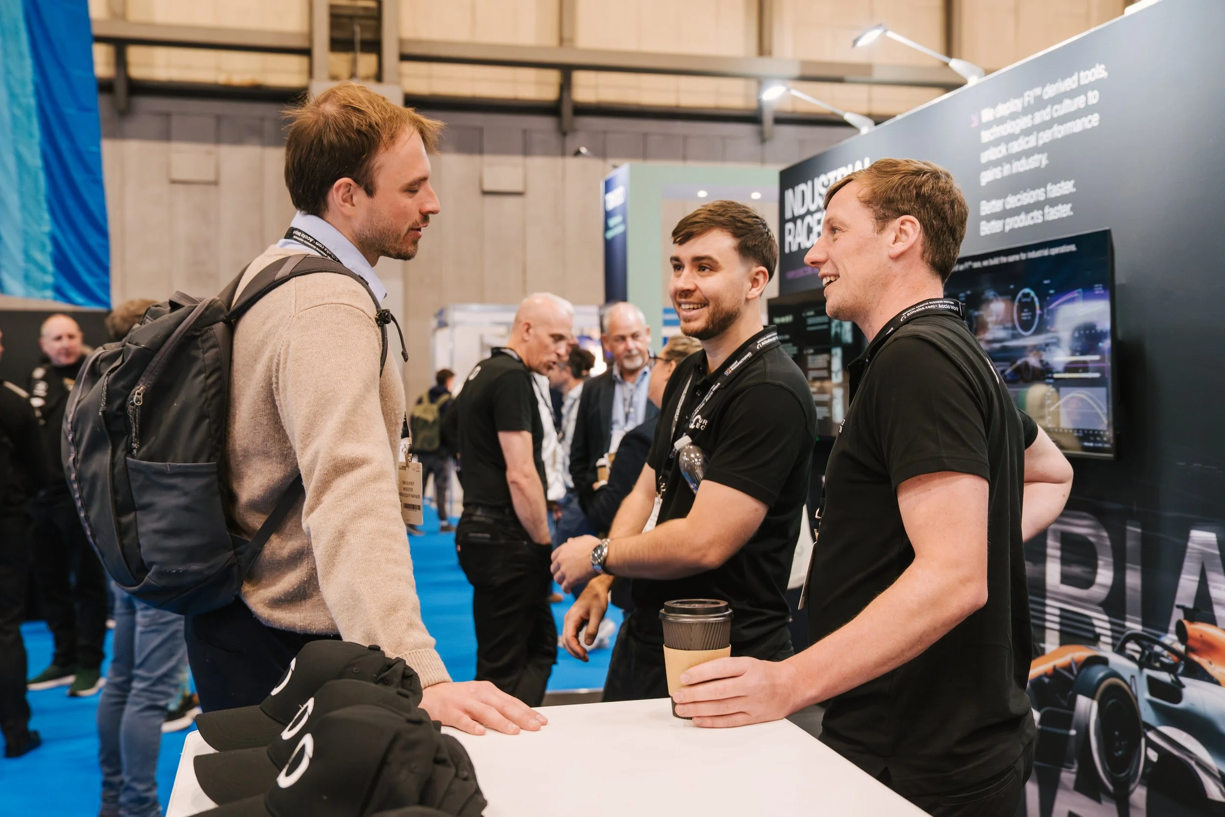 Two men are engaged in conversation at a trade show booth, with people in the background. One man carries a backpack and the other holds a coffee cup.