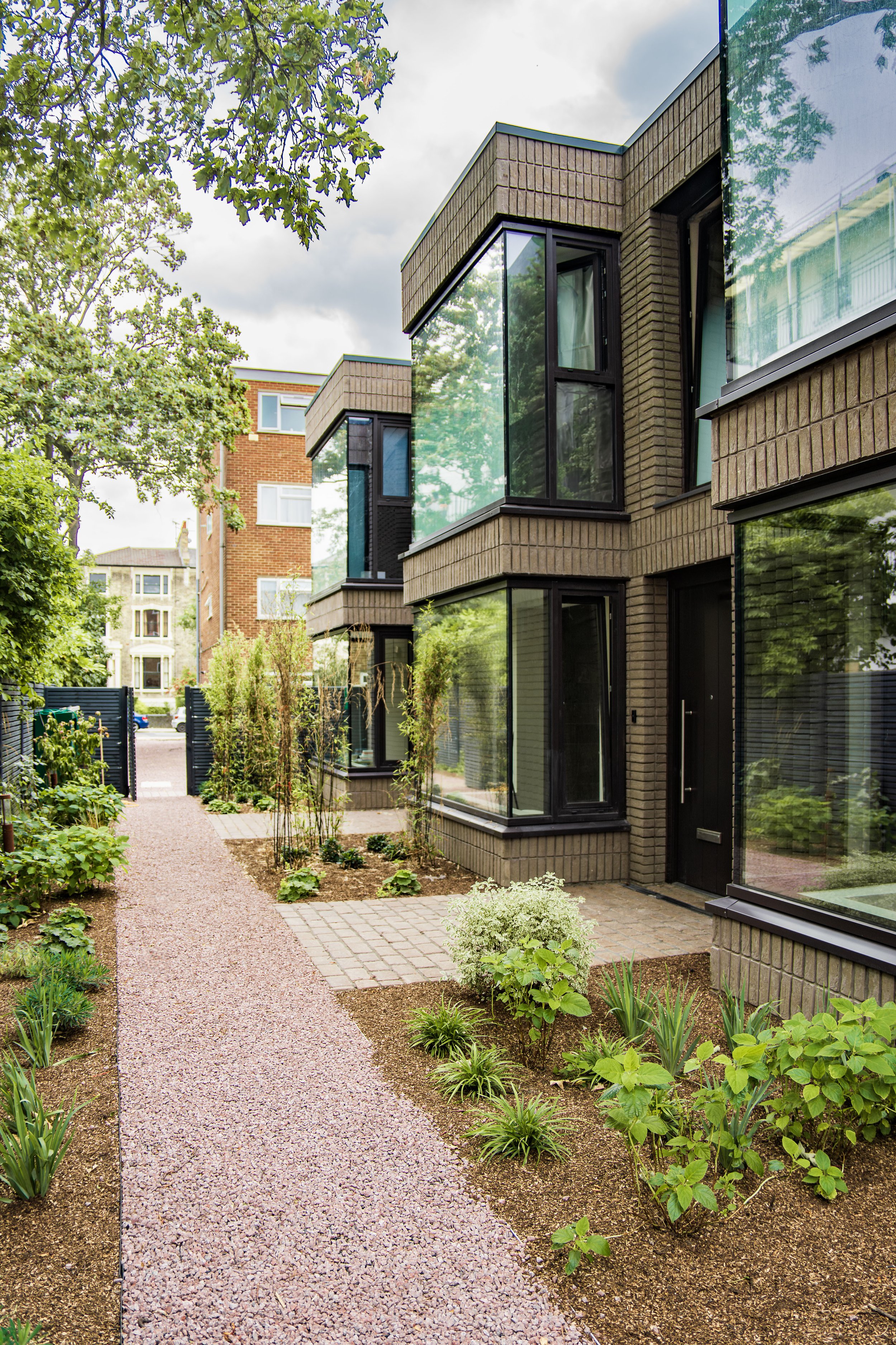 A modern urban building with large glass windows and brown brick exterior, surrounded by a well-maintained garden with a gravel path, small shrubs, and trees.