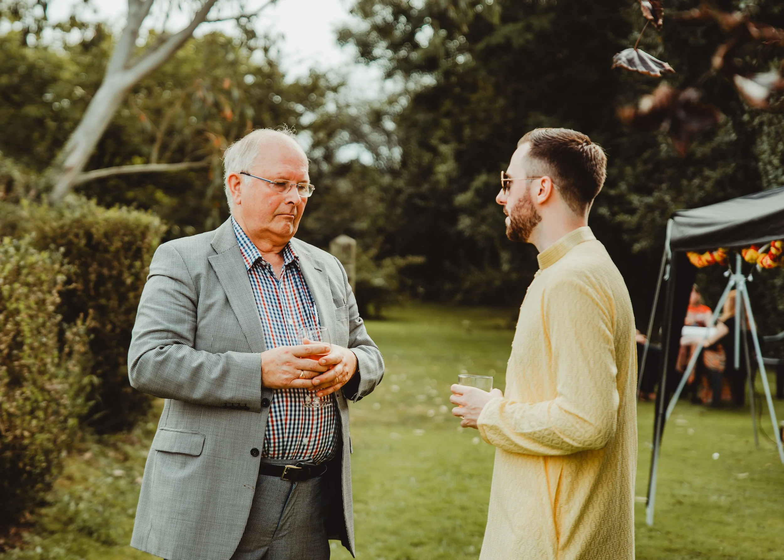 Two men are outdoors, one older with glasses and a gray blazer holding a drink, the other younger with sunglasses and a yellow sweater also holding a drink, engaged in conversation at a gathering.