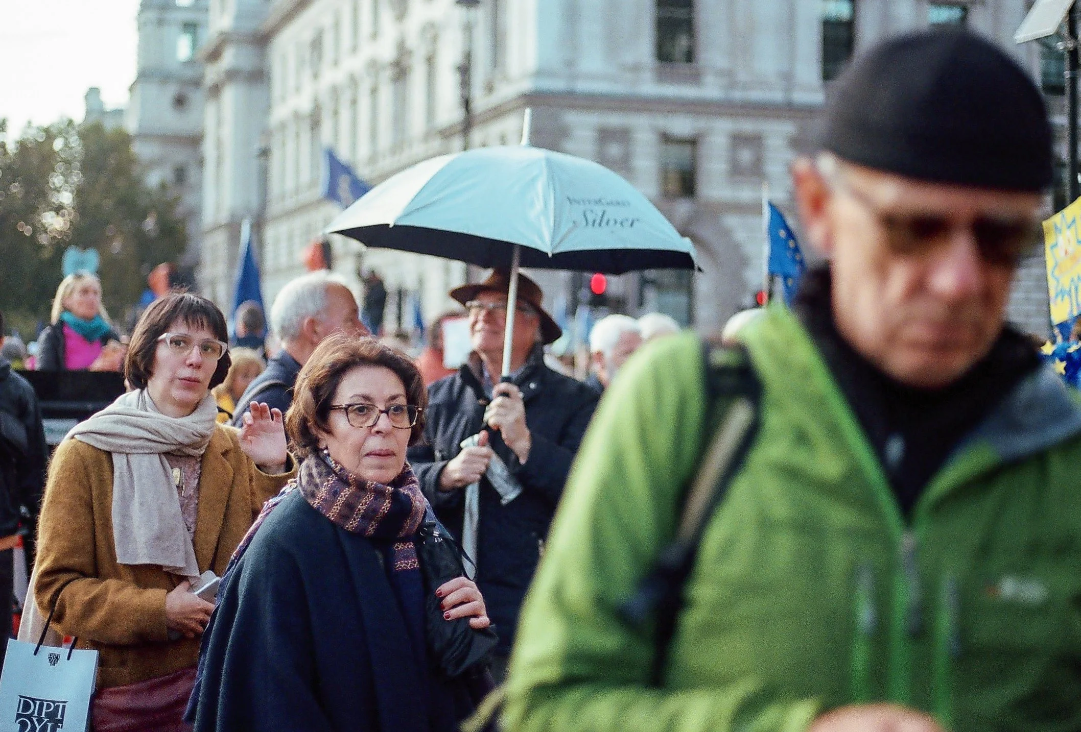 Crowd of people participating in a protest or rally on a city street, some holding signs and umbrellas, with historic buildings in the background.
