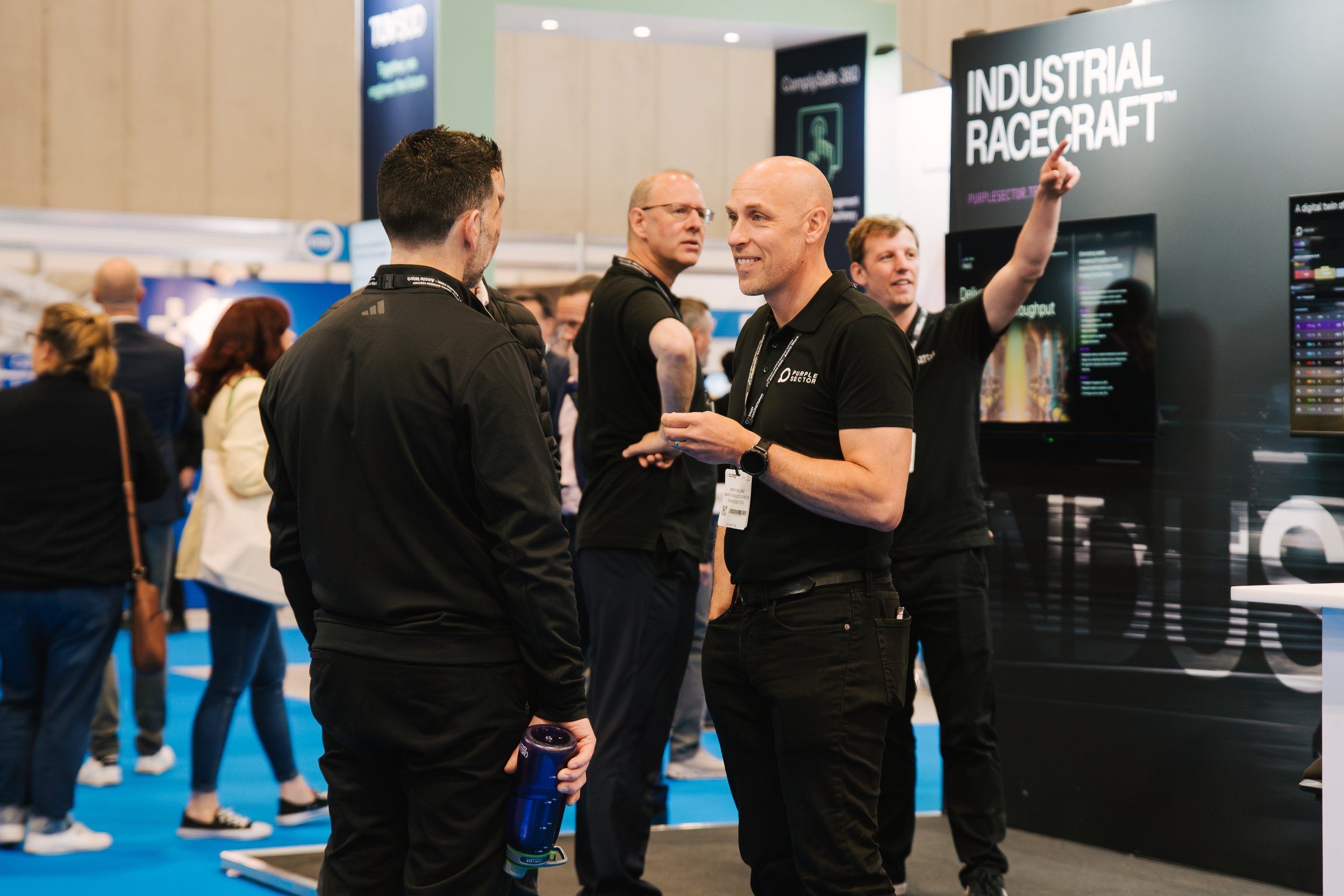 Three men are in conversation at an event booth with the signage 'Industrial Racecraft'. One man with a shaved head is smiling and gesturing, while another man in a black jacket holds a water bottle. A third man with glasses looks on in the backgroun
