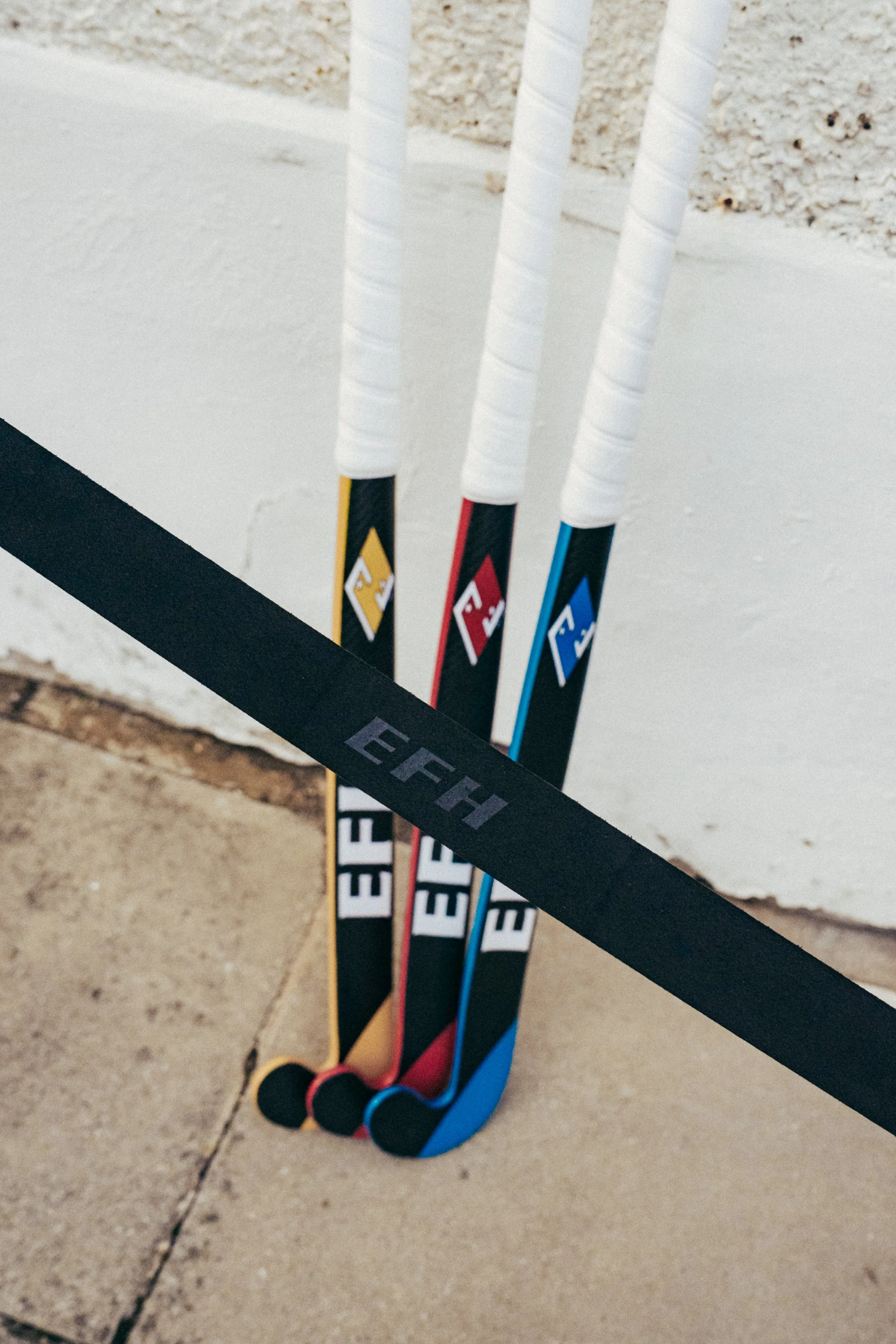 A set of three hockey sticks with different colored handles and white grips, leaning against a black fence on a concrete surface, with a textured wall in the background.