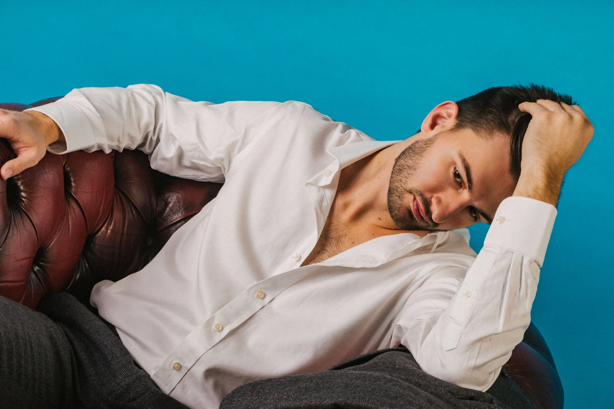 A young man with dark hair and a beard lying on a burgundy leather couch, resting his head on his hand, against a blue background.