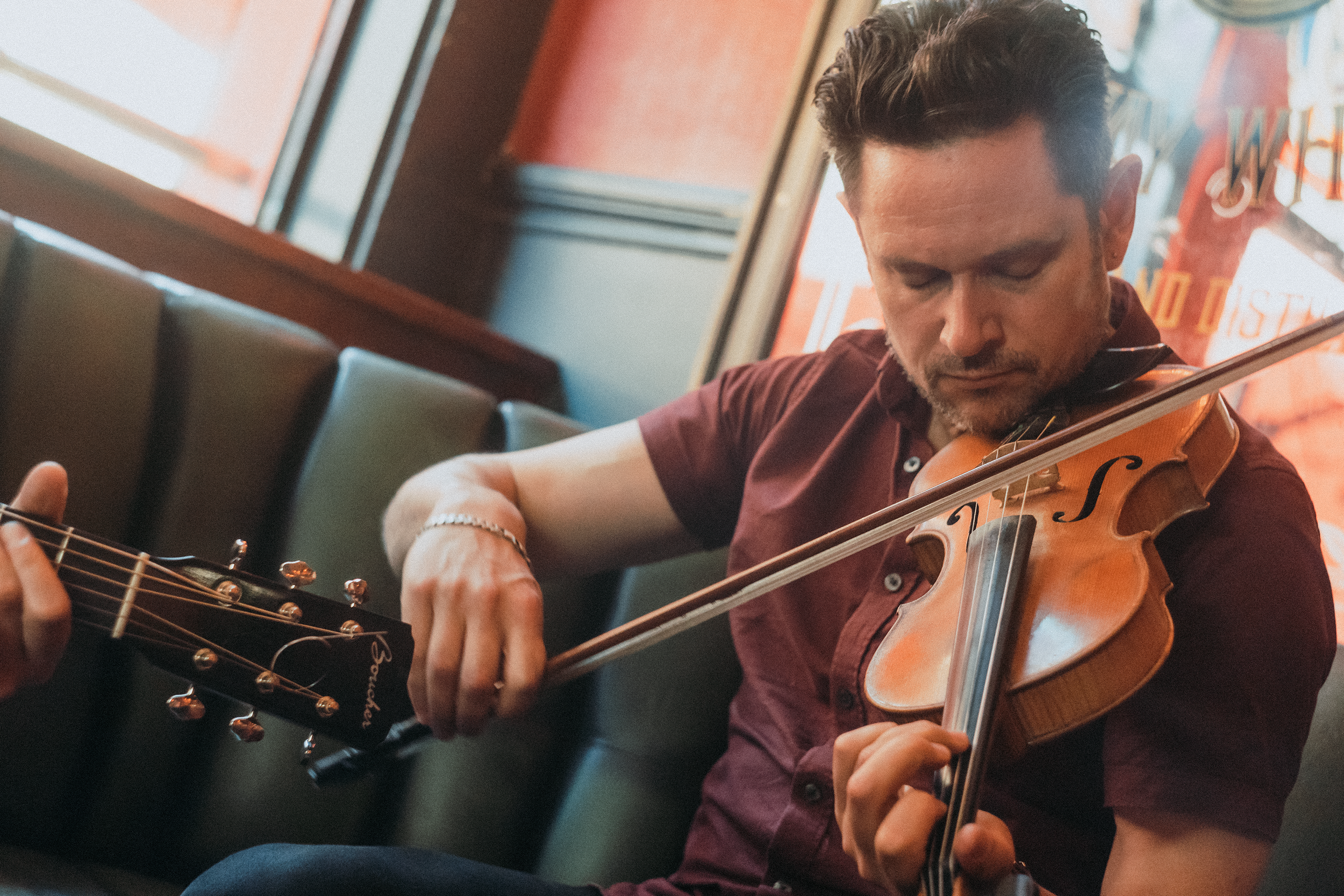 A man with dark hair and a beard playing a violin in a casual setting, possibly a cafe or restaurant, with wall art and a window in the background.
