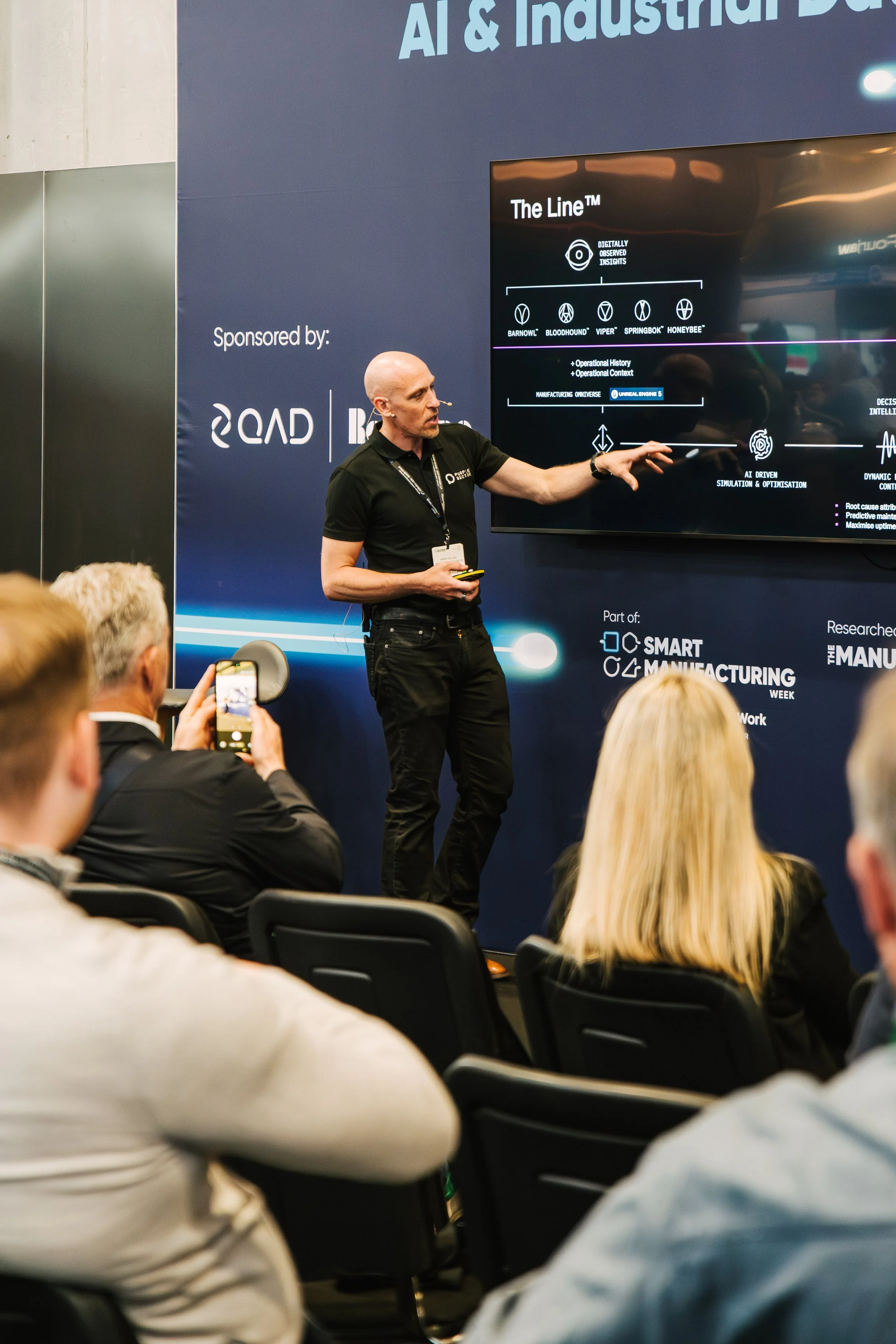 A man giving a presentation at a conference about AI and industrial automation, with a large screen displaying a slide titled 'The Line™' and audience members taking photos.