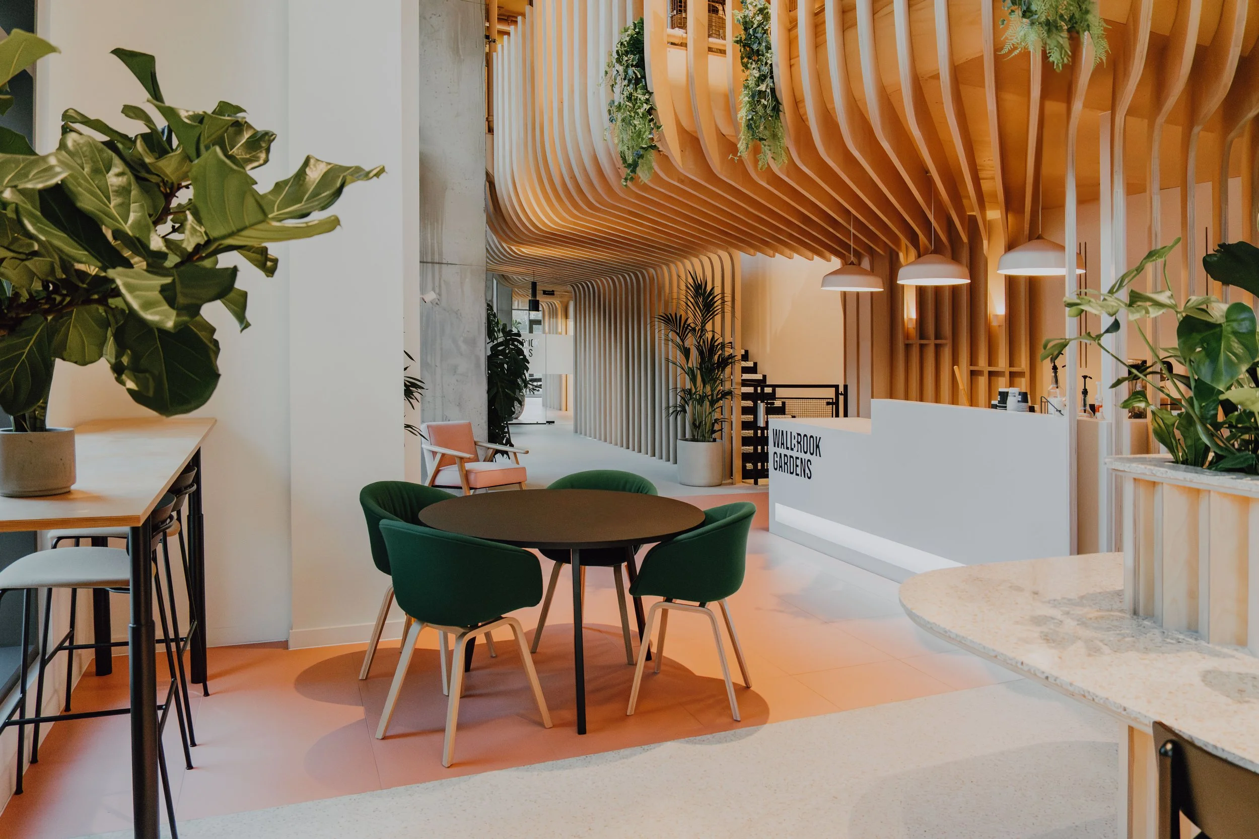 Modern indoor space with light wood architectural features, green plants, green and pink chairs, white terrazzo counters, and pendant lighting.