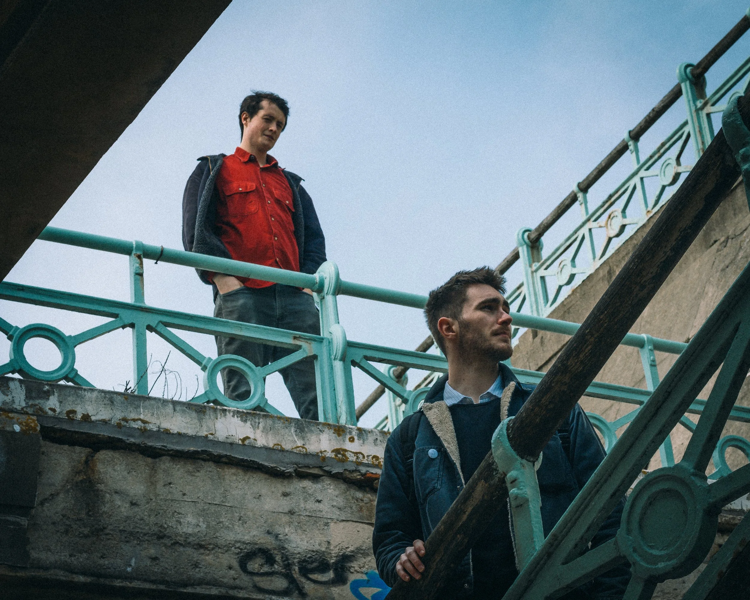 Two young men on a balcony with a teal decorative railing, one standing above the other, looking in different directions, with a cloudy sky background.