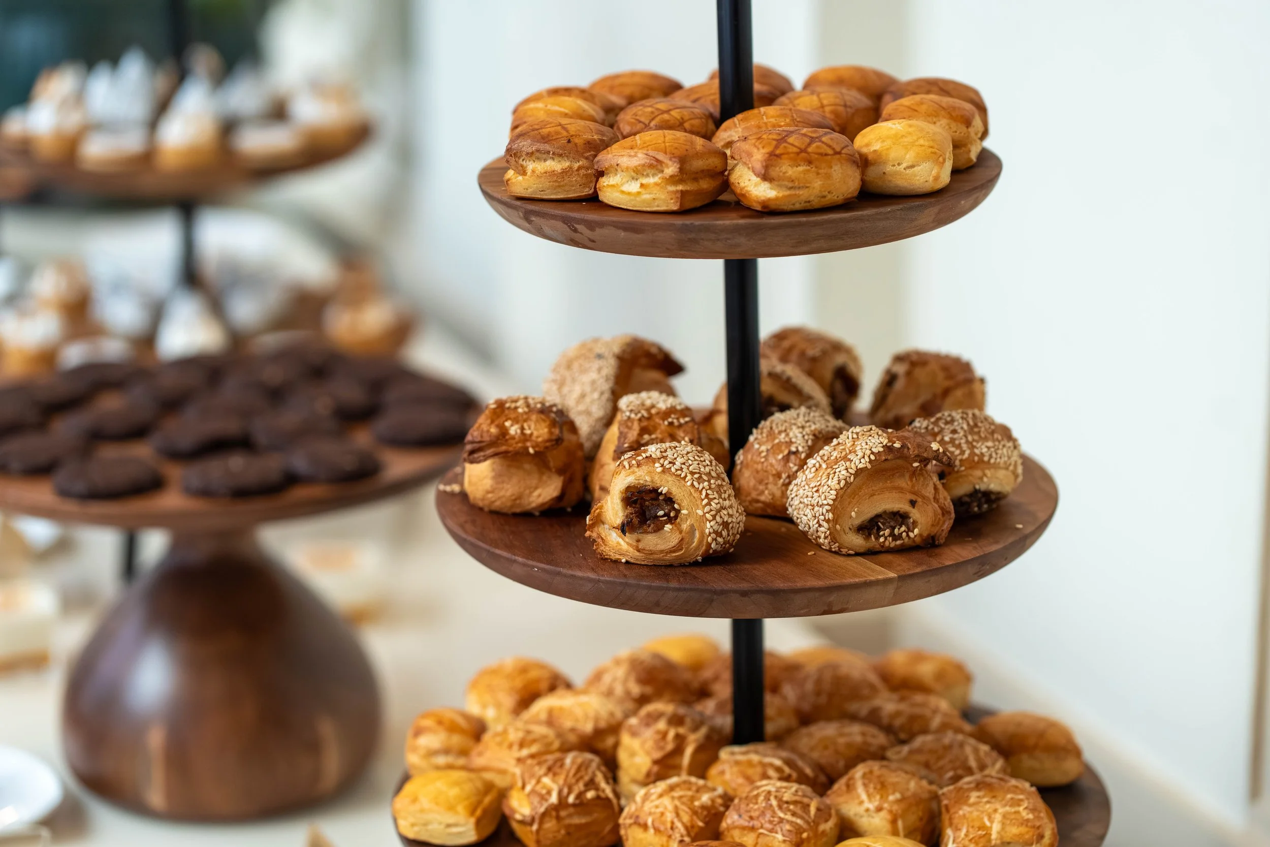 Three-tiered wooden tray filled with assorted pastries, including cream-filled éclairs with sesame seeds, chocolate-covered pastries, and other baked goods, at a dessert table.