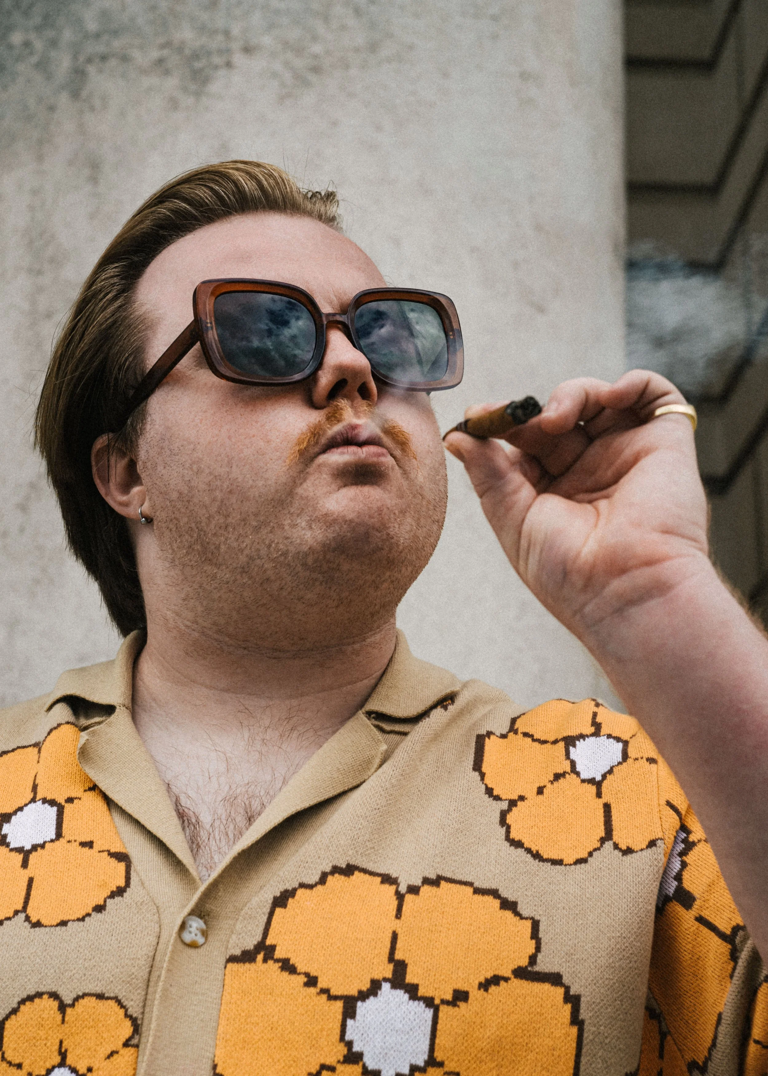 A person with light skin wearing large dark sunglasses, a beige shirt with a colorful tiger print, and smoking a cigar, standing against a concrete wall.