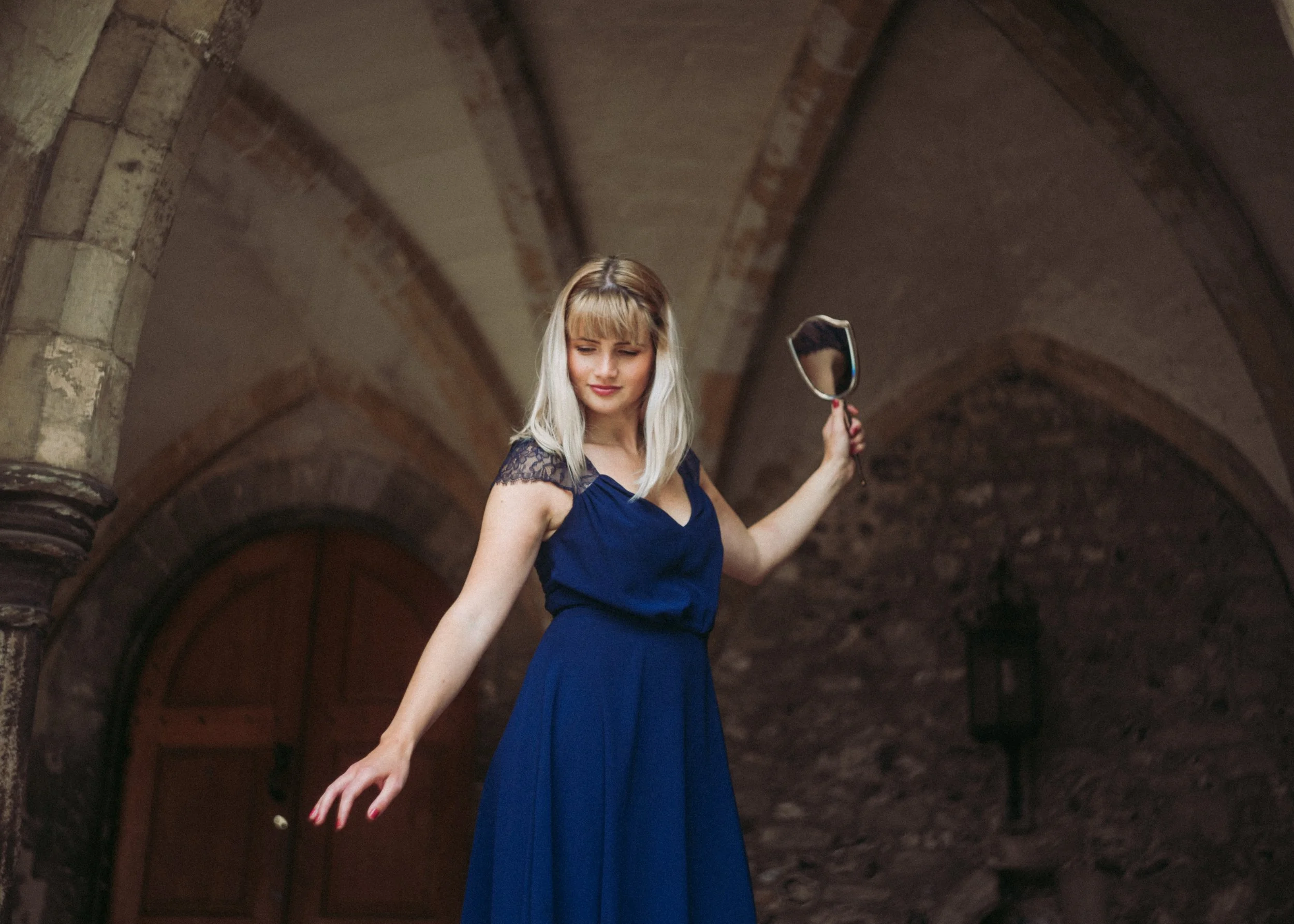 A woman in a dark blue dress holding a mirror in an underground stone corridor.