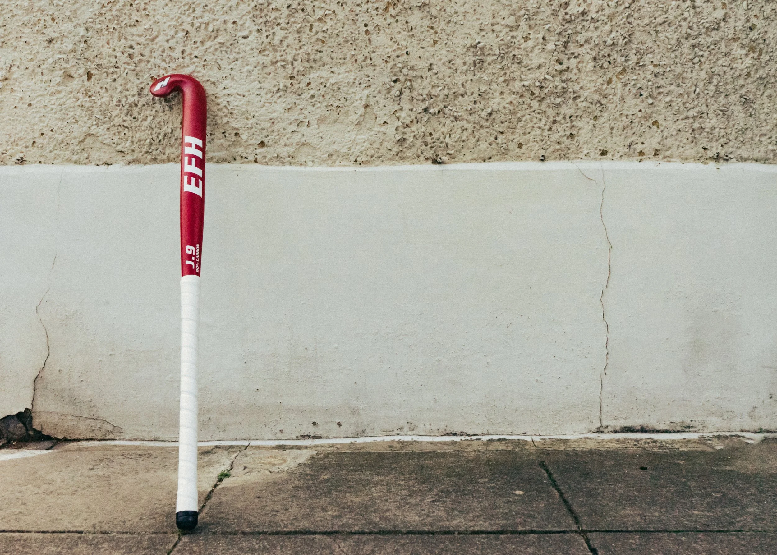 A red and white baseball bat with the word 'DEEP' on it leaning against a beige wall with cracks at the bottom, on a concrete ground.