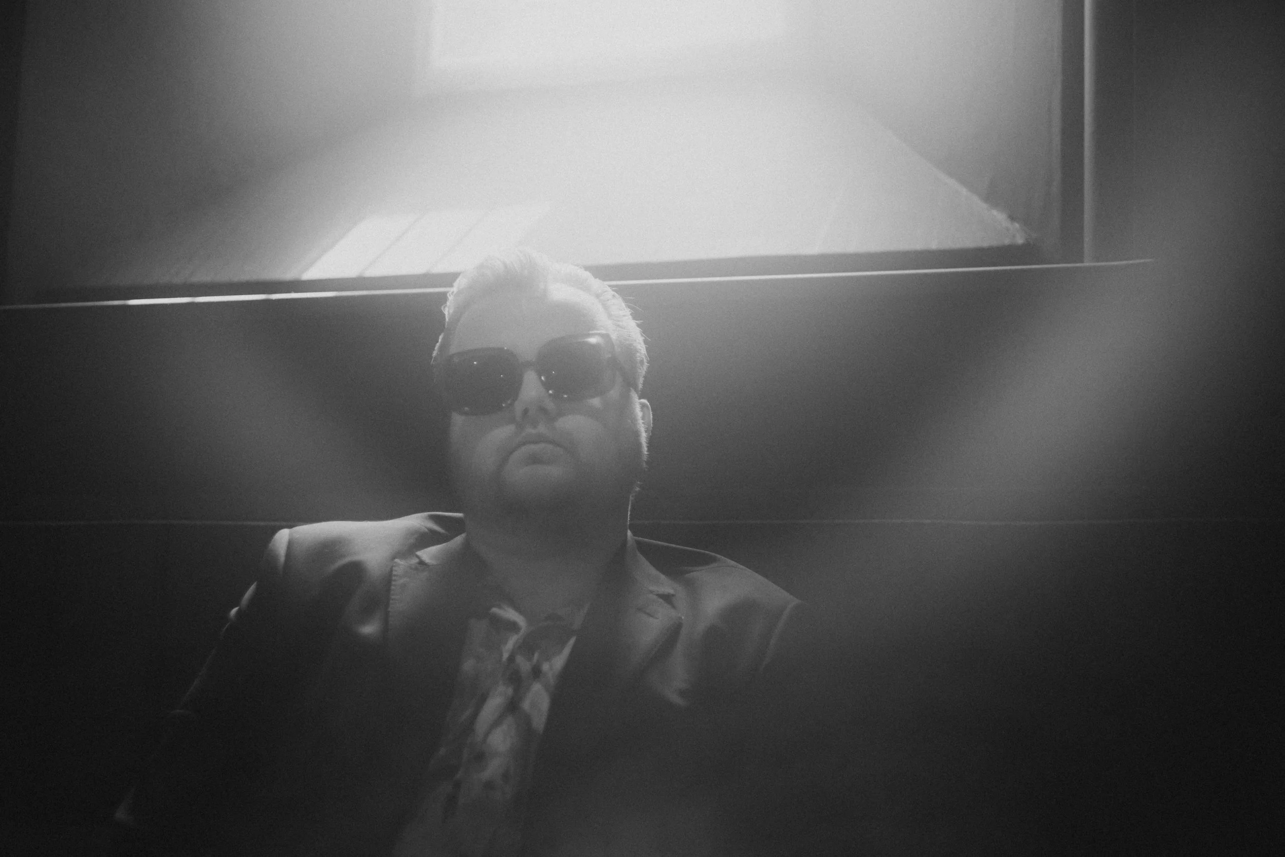 Black and white photo of a person with short hair and sunglasses, looking down at the camera, with their lower face and chest visible.