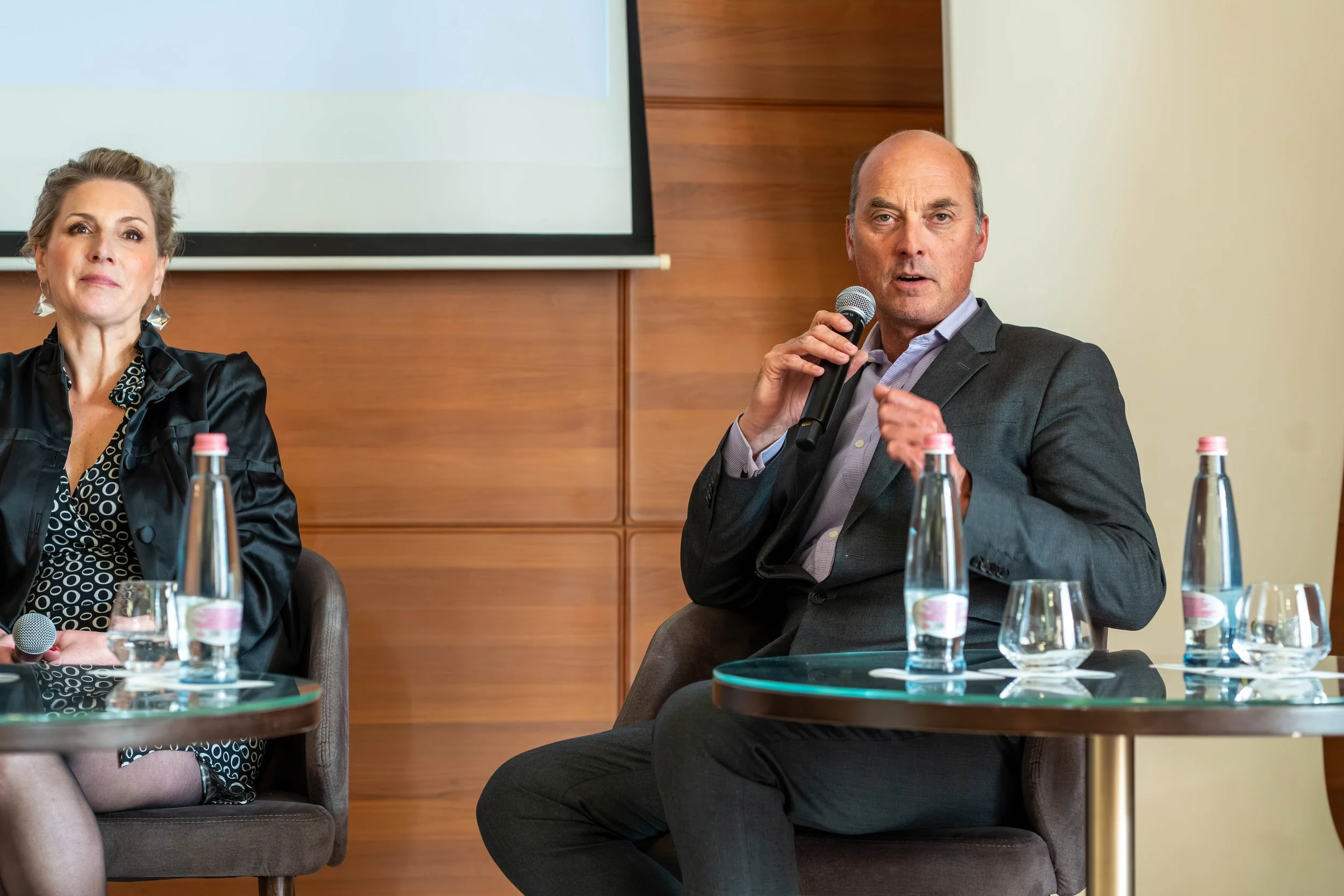 A man in a gray suit and a woman in a black outfit sitting at a conference table with water bottles and glasses, participating in a discussion or presentation in a professional setting.