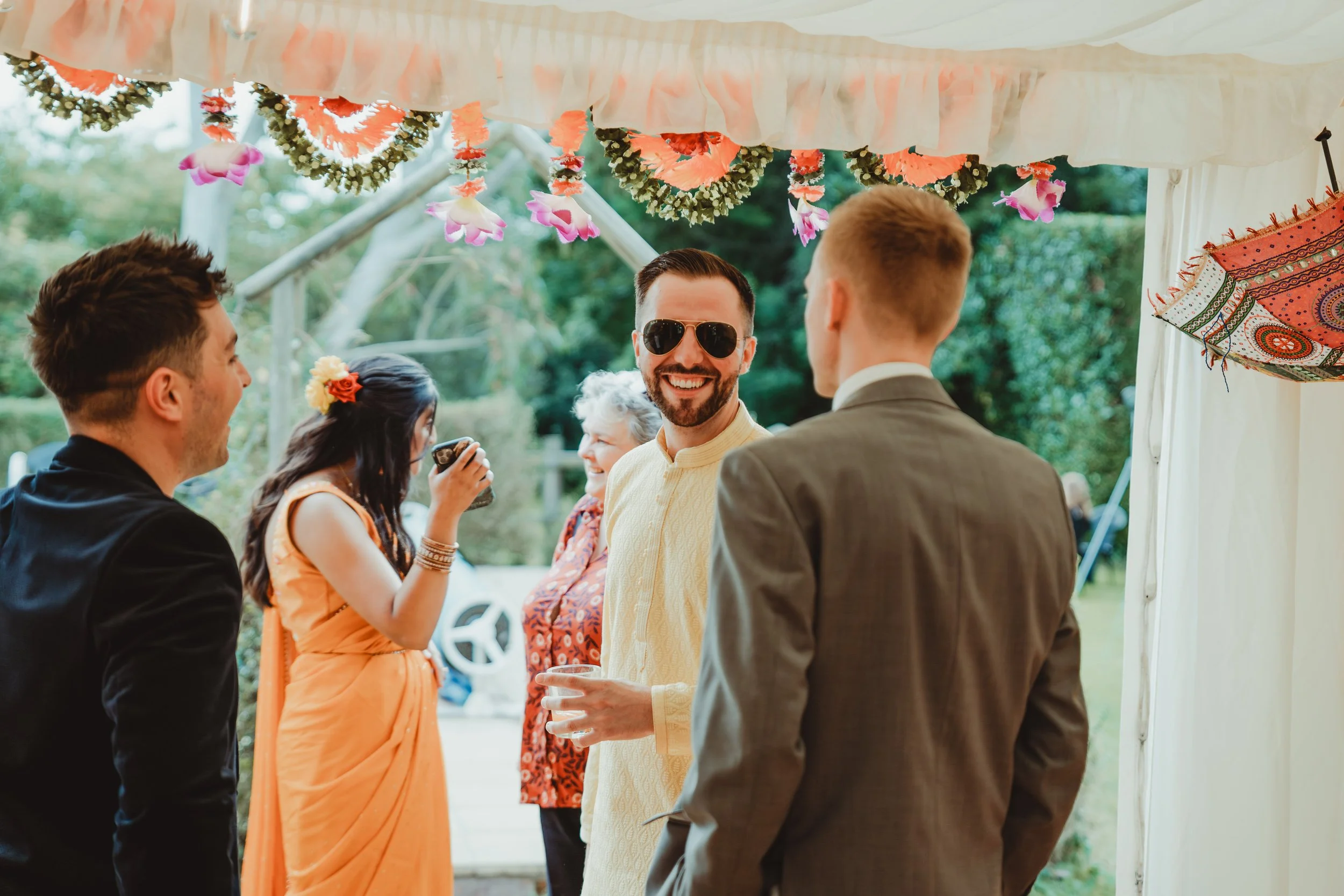 A group of people at an outdoor celebration under a decorated tent, engaging in conversation, with some smiling and one person holding a drink.