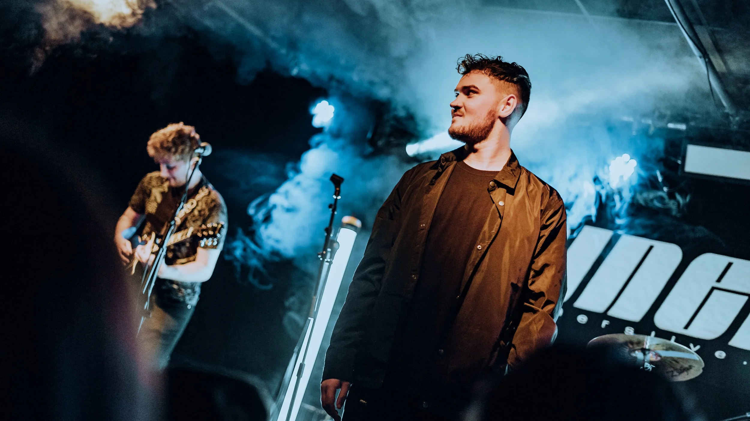 Two musicians perform on stage at a concert, with one playing a guitar and the other standing nearby, illuminated by stage lights.