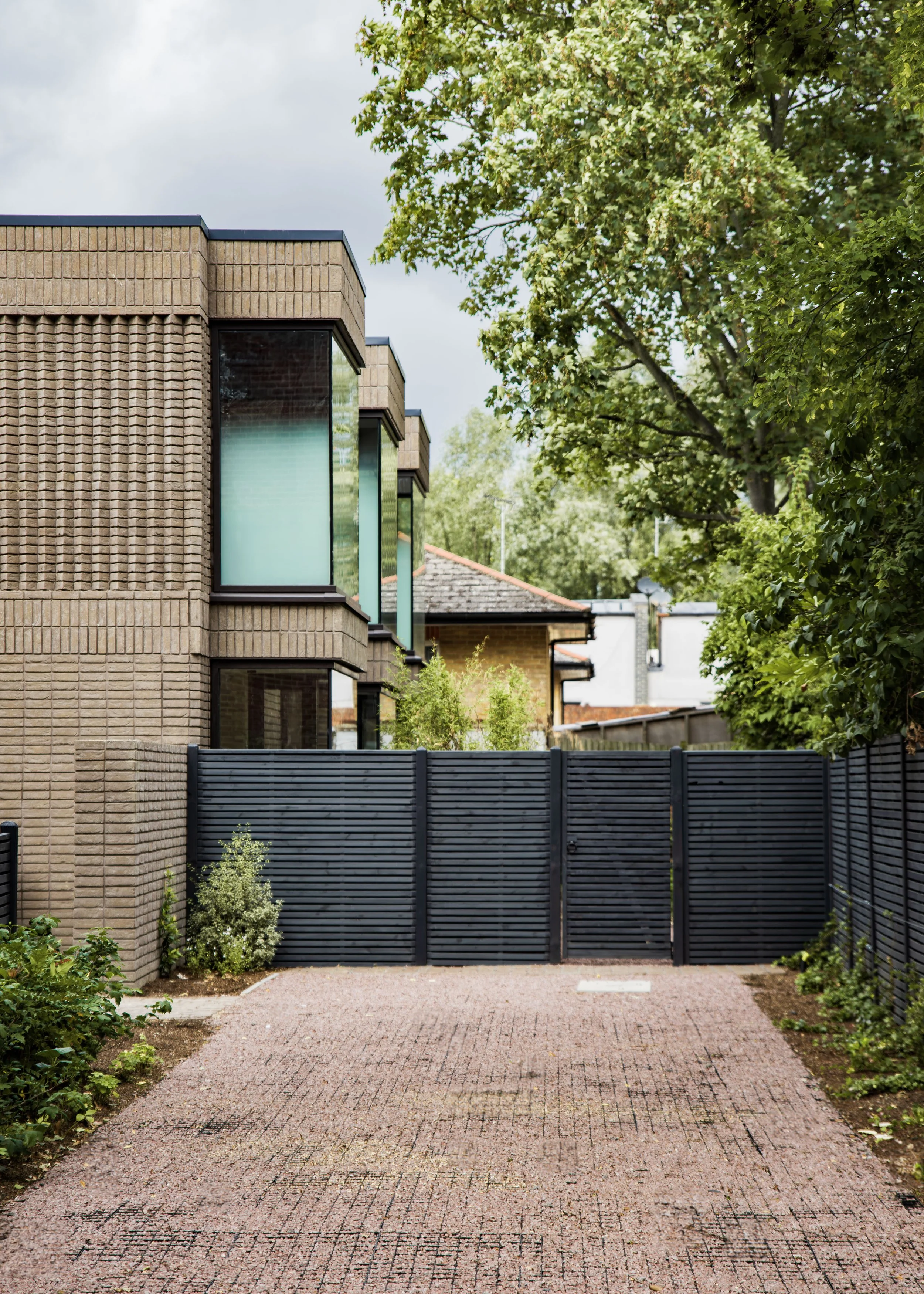 A modern building with large glass windows and a brick exterior, surrounded by trees and greenery, with a paved driveway in the foreground and a black fence.