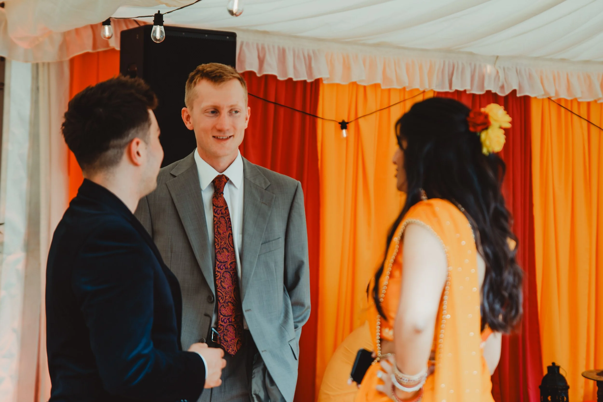A wedding ceremony with a groom and a bride standing in front of an officiant, with orange and yellow curtains in the background and hanging light bulbs from the ceiling.