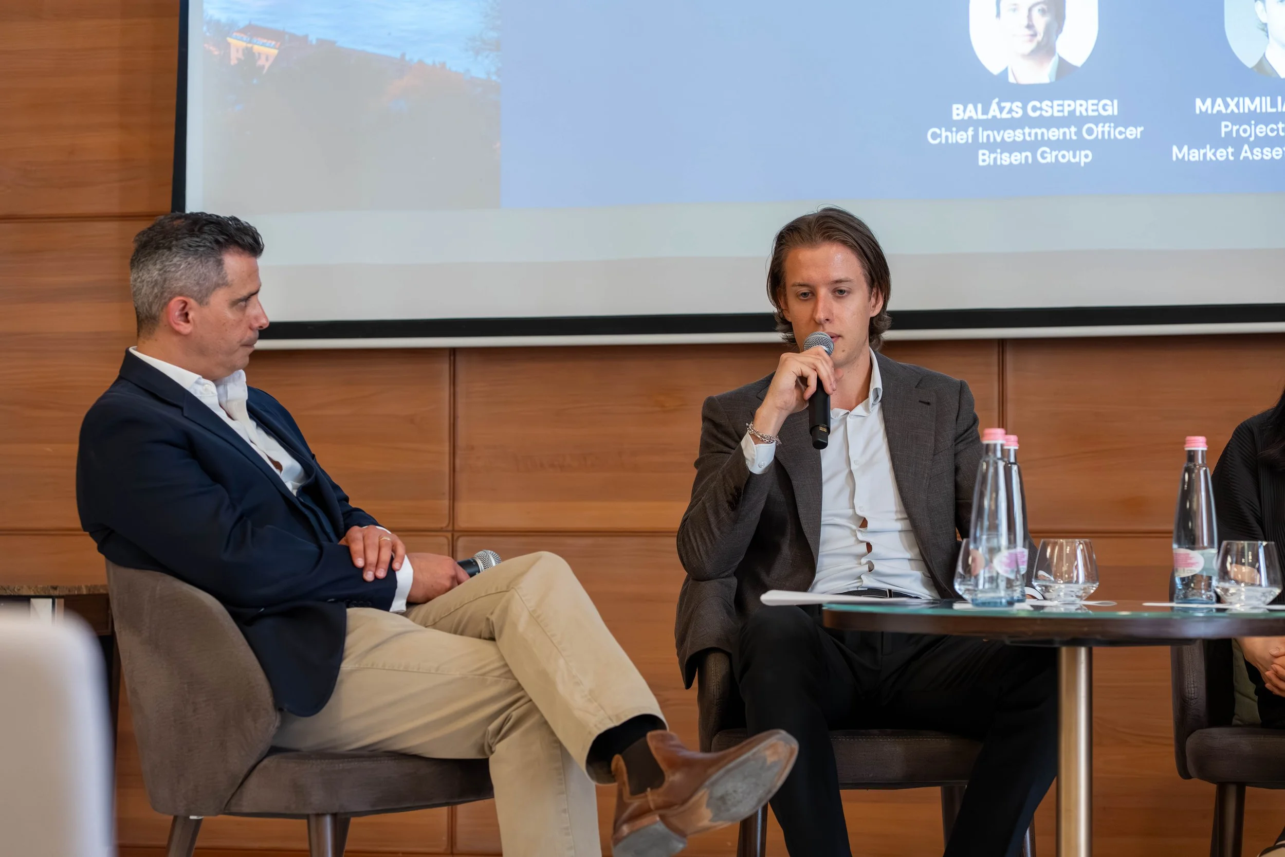 Two men sitting on chairs at a conference table, one is speaking into a microphone, with a presentation slide in the background.
