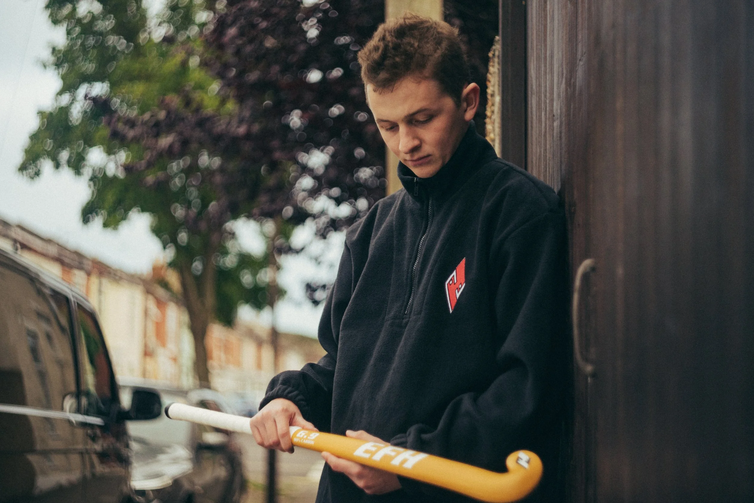 A young man with short brown hair, wearing a black jacket, is standing outside near a black fence or wall. He is holding a yellow hockey stick with white and black details, and appears to be looking down at it thoughtfully. There are cars parked on t