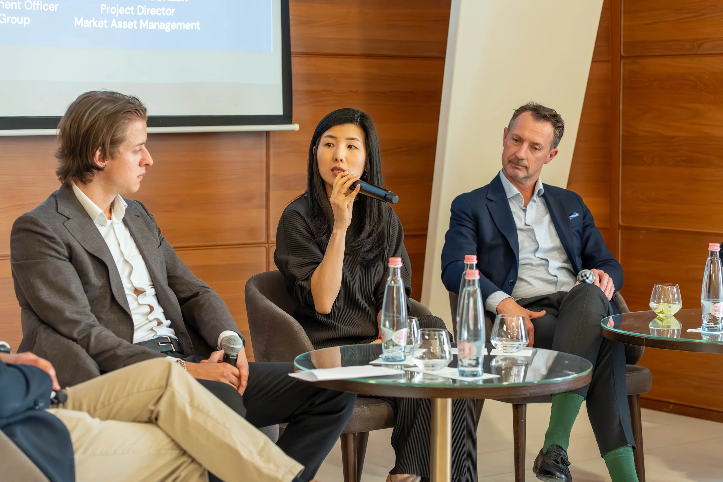Three people sitting at a conference table during a panel discussion, with bottles of water and glasses in front of them. The woman in the center is speaking into a microphone. A presentation slide is visible in the background.