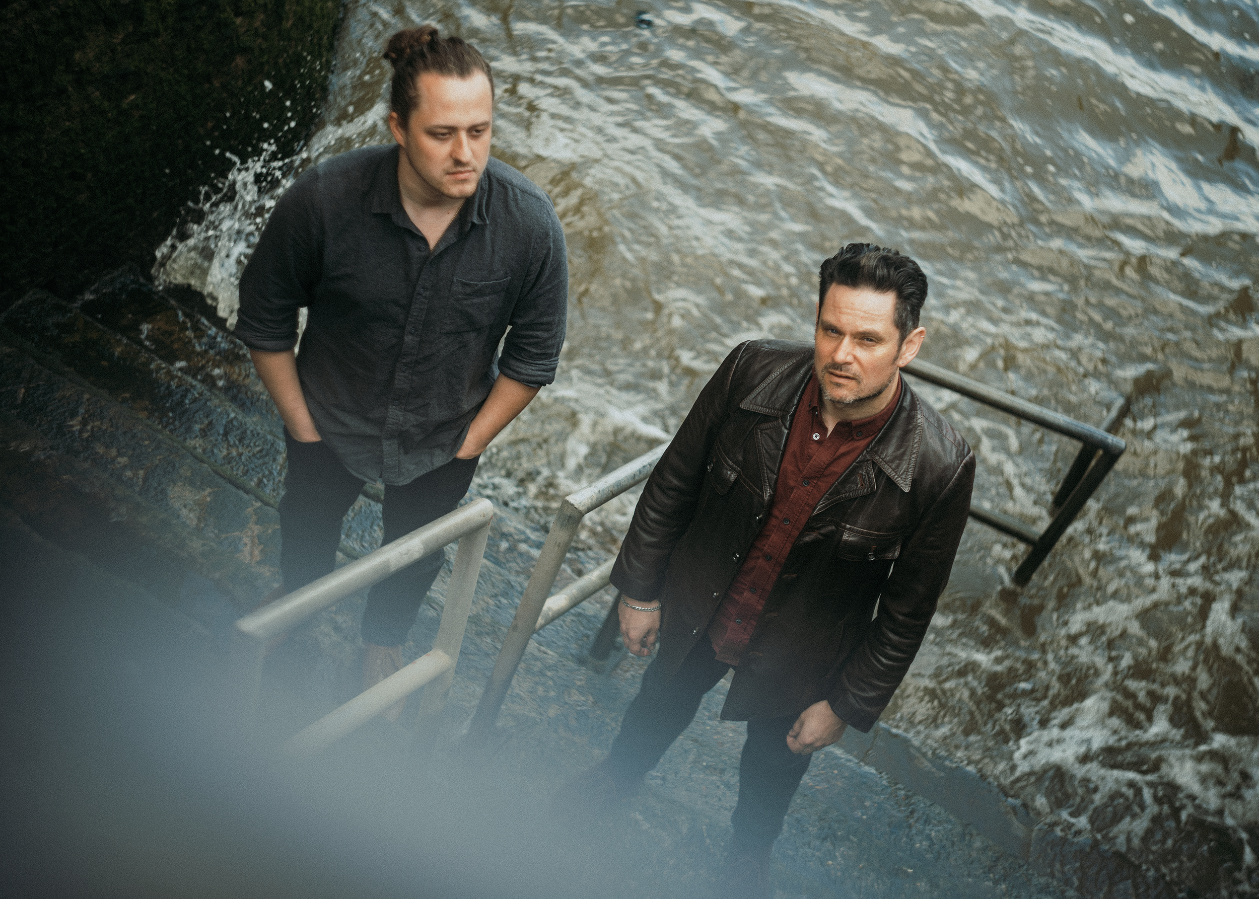 Two men walking up a staircase by a body of water, one wearing a dark leather jacket and the other in a dark shirt with rolled-up sleeves.