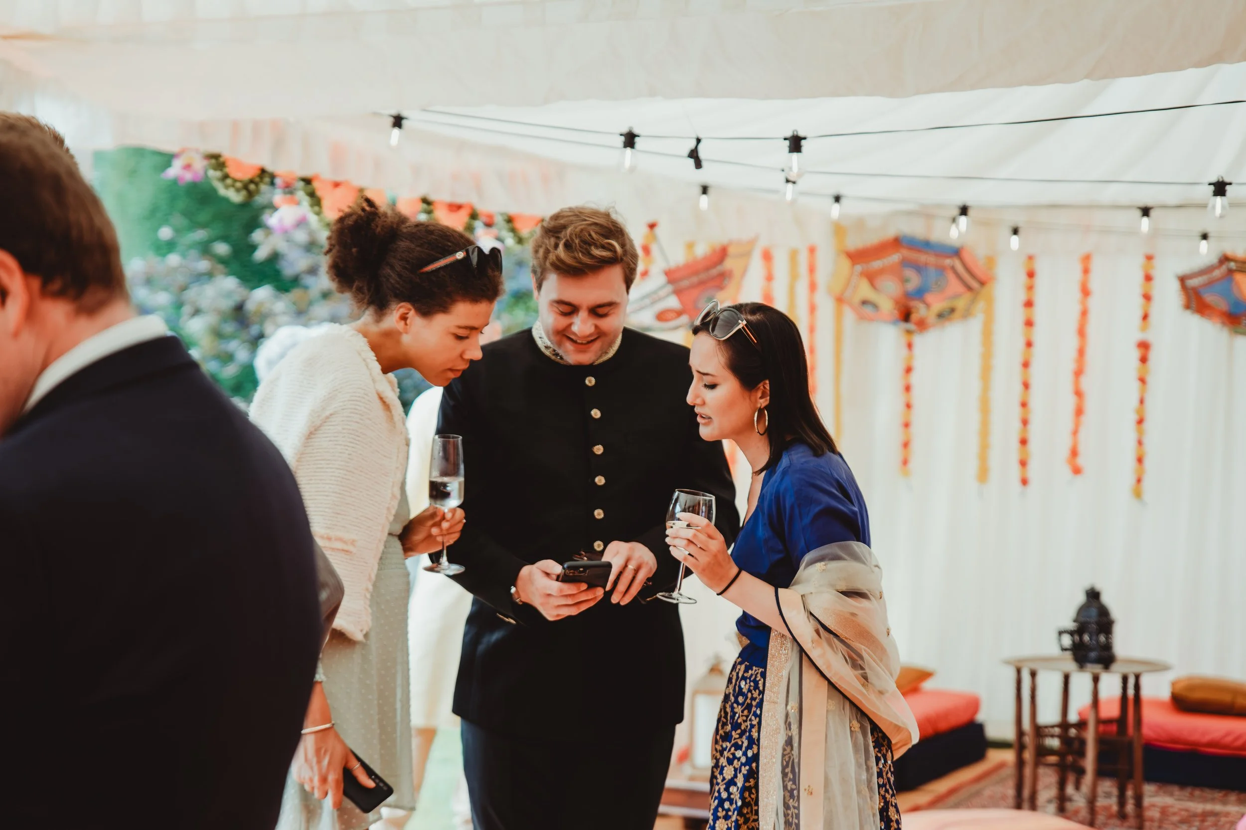 Group of people at a festive event, looking at a smartphone together, with colorful decorations and string lights in the background.