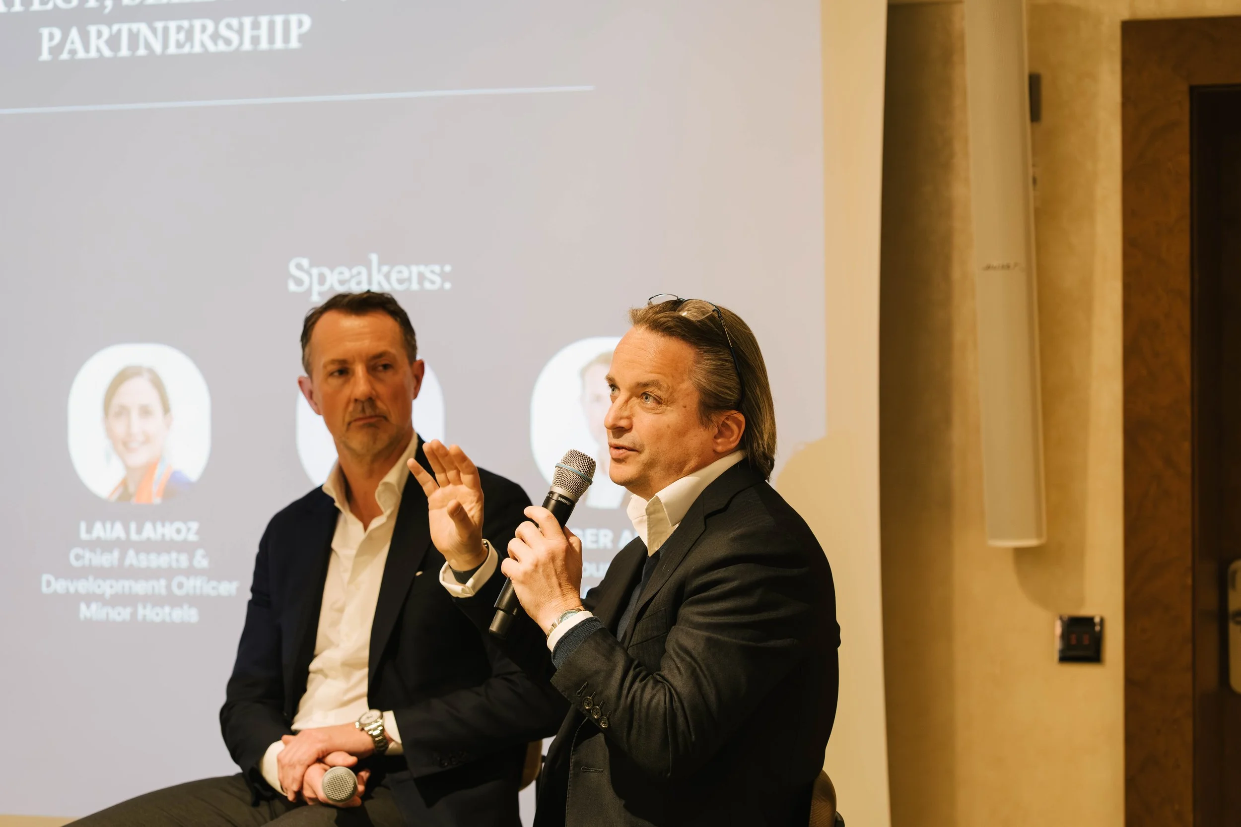 Two men speaking at an event, one holding a microphone, with a presentation slide in the background.