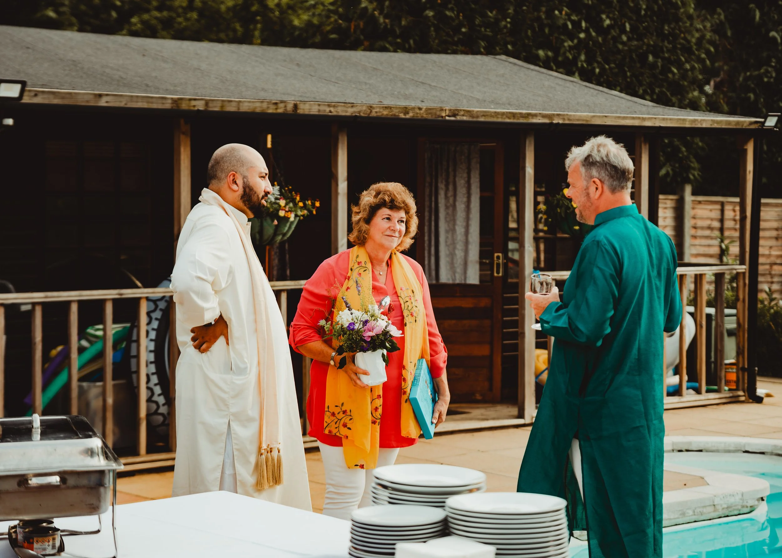 Three people are standing outdoors near a house, engaging in conversation. The woman in the center is holding a bouquet of flowers and a book. The man on the right is smiling and holding a glass, while another man on the left listens attentively. The