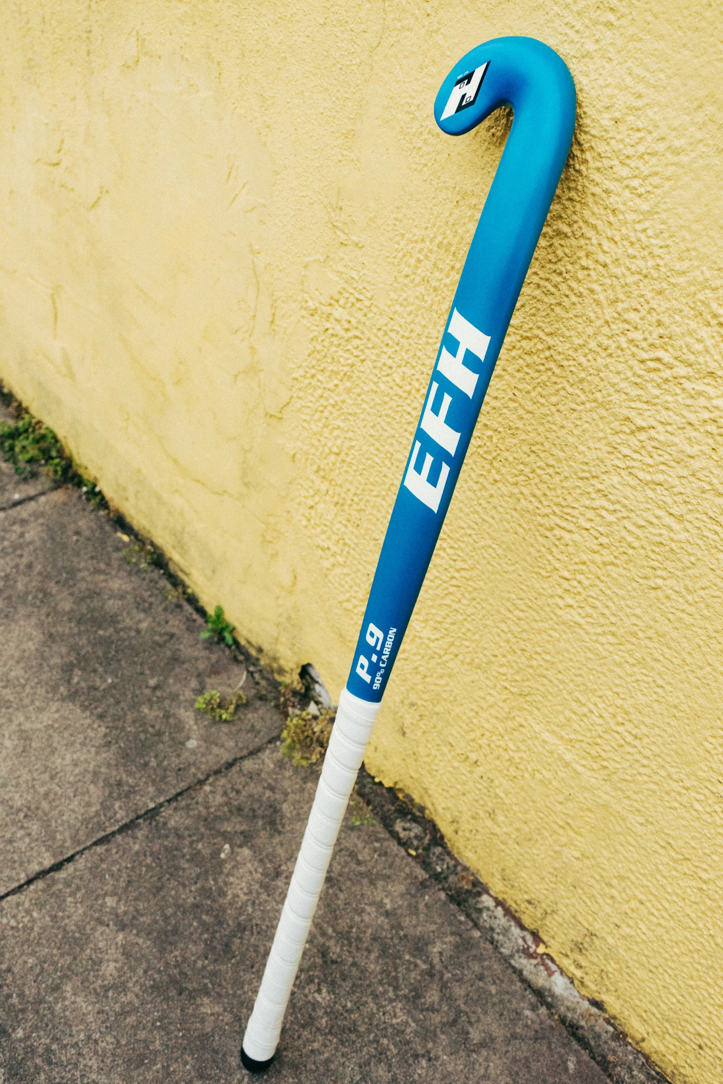 Blue hockey stick with white handle leaning against a yellow textured wall on a sidewalk.