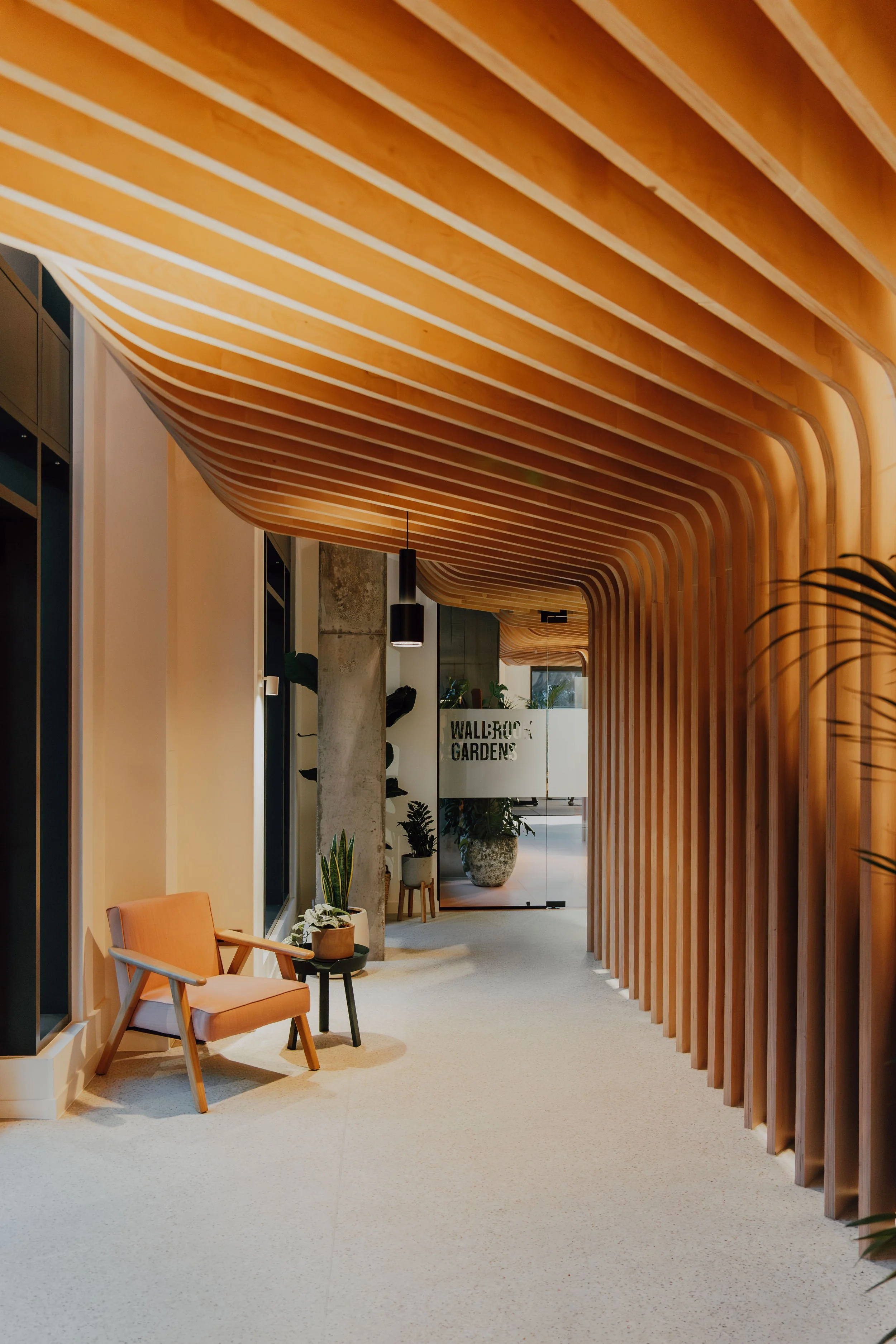 Modern indoor corridor with curved wooden slat ceiling, beige carpet, orange armchair and small side table with plant, potted plants and glass door in background.