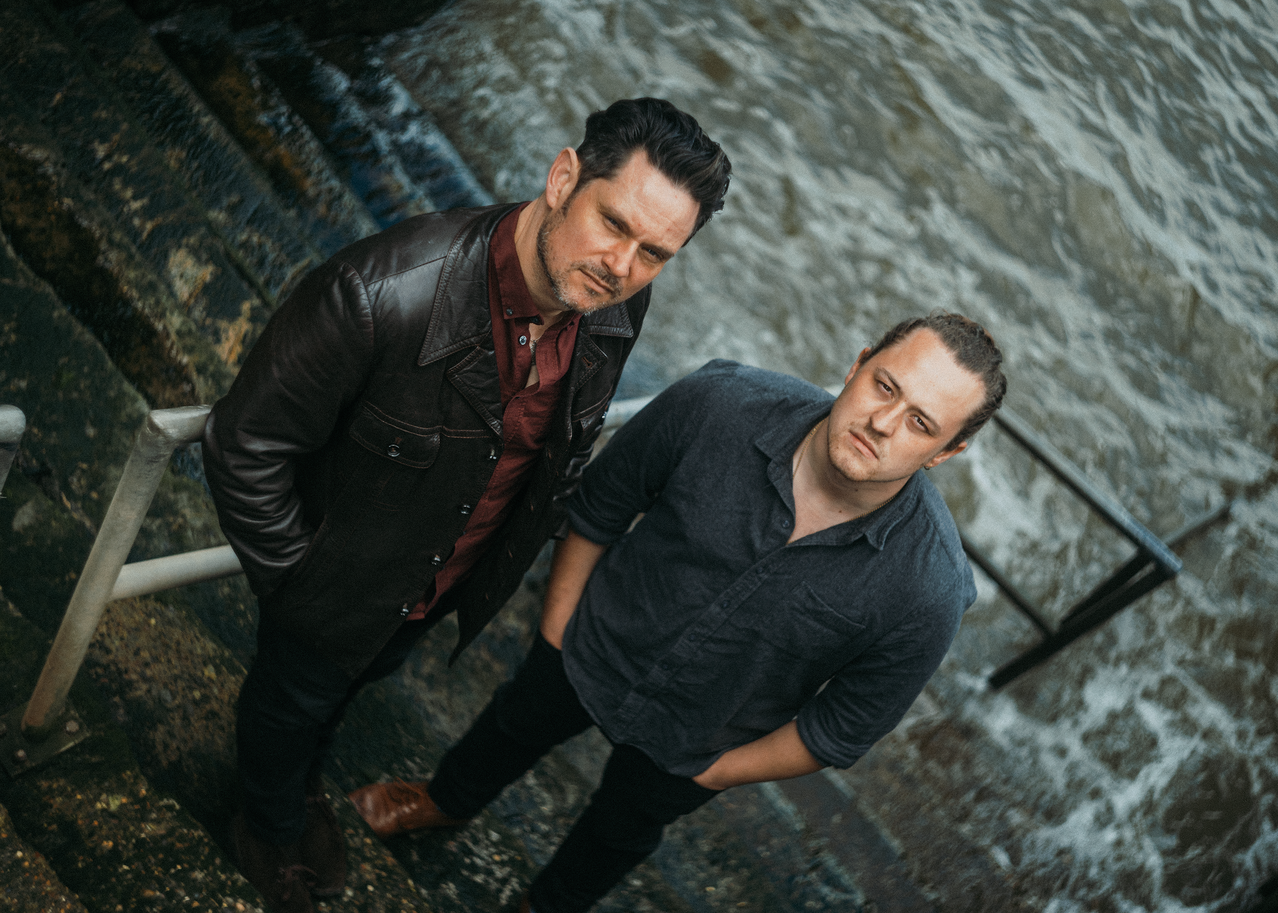 Two men stand on a rocky, wet surface near a body of water, looking at the camera with serious expressions.