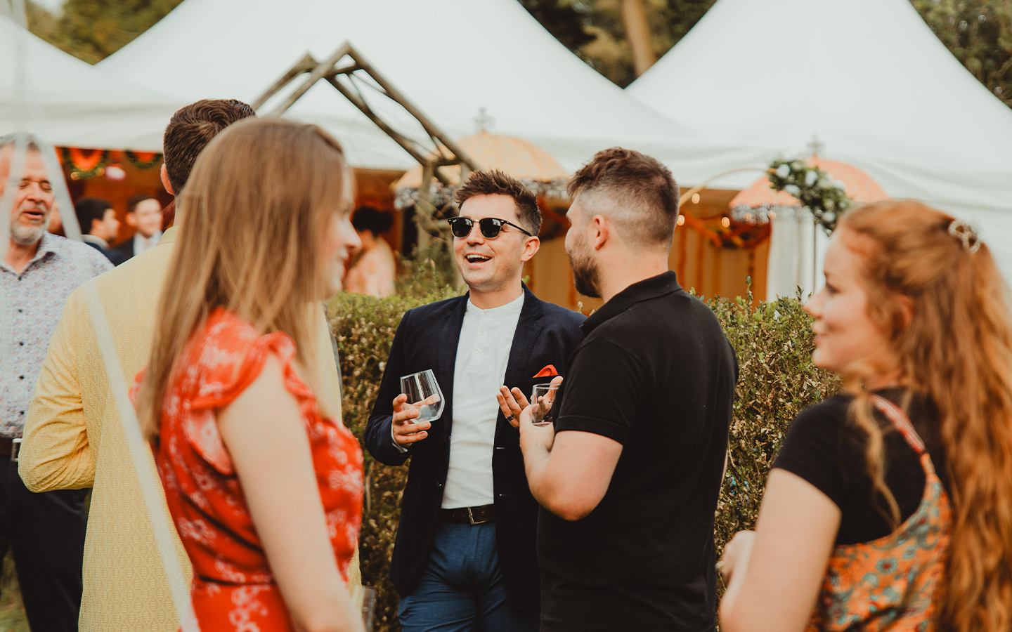 A group of people socializing outdoors at a gathering, with white tents and decorations visible in the background. They are engaged in conversation, holding drinks, and dressed in semi-formal attire.