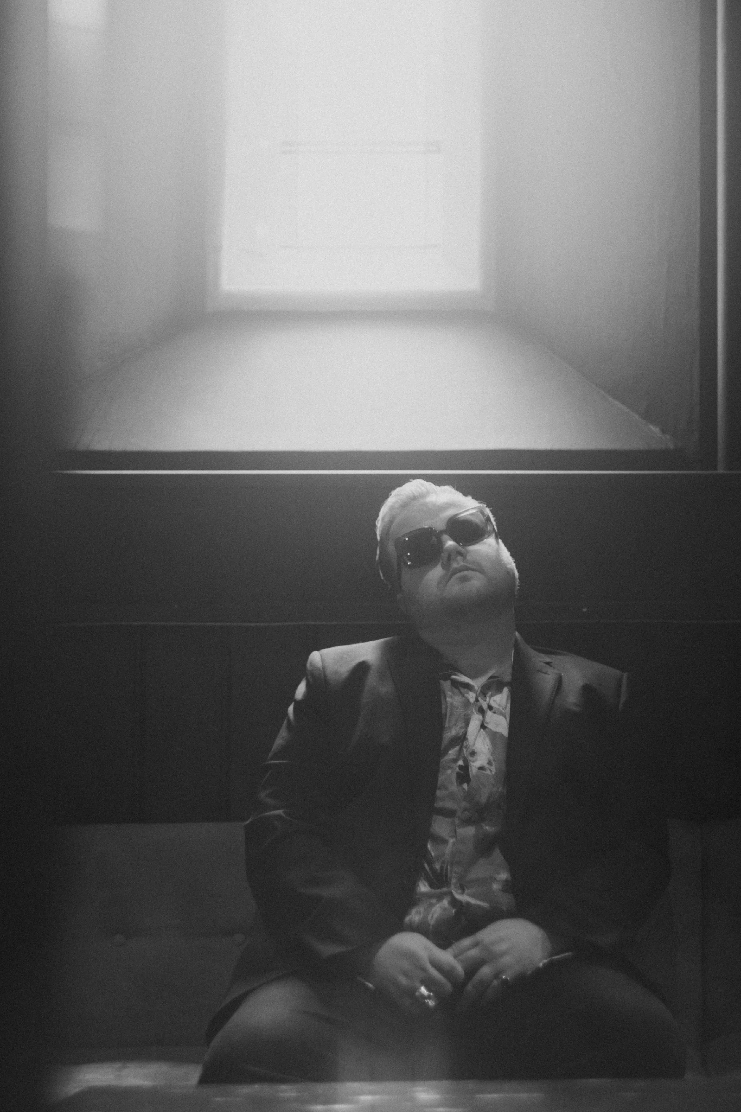 A man wearing sunglasses and a suit is sitting in an elevator taking a selfie. The elevator has a plain ceiling with a bright rectangular light and dark walls.