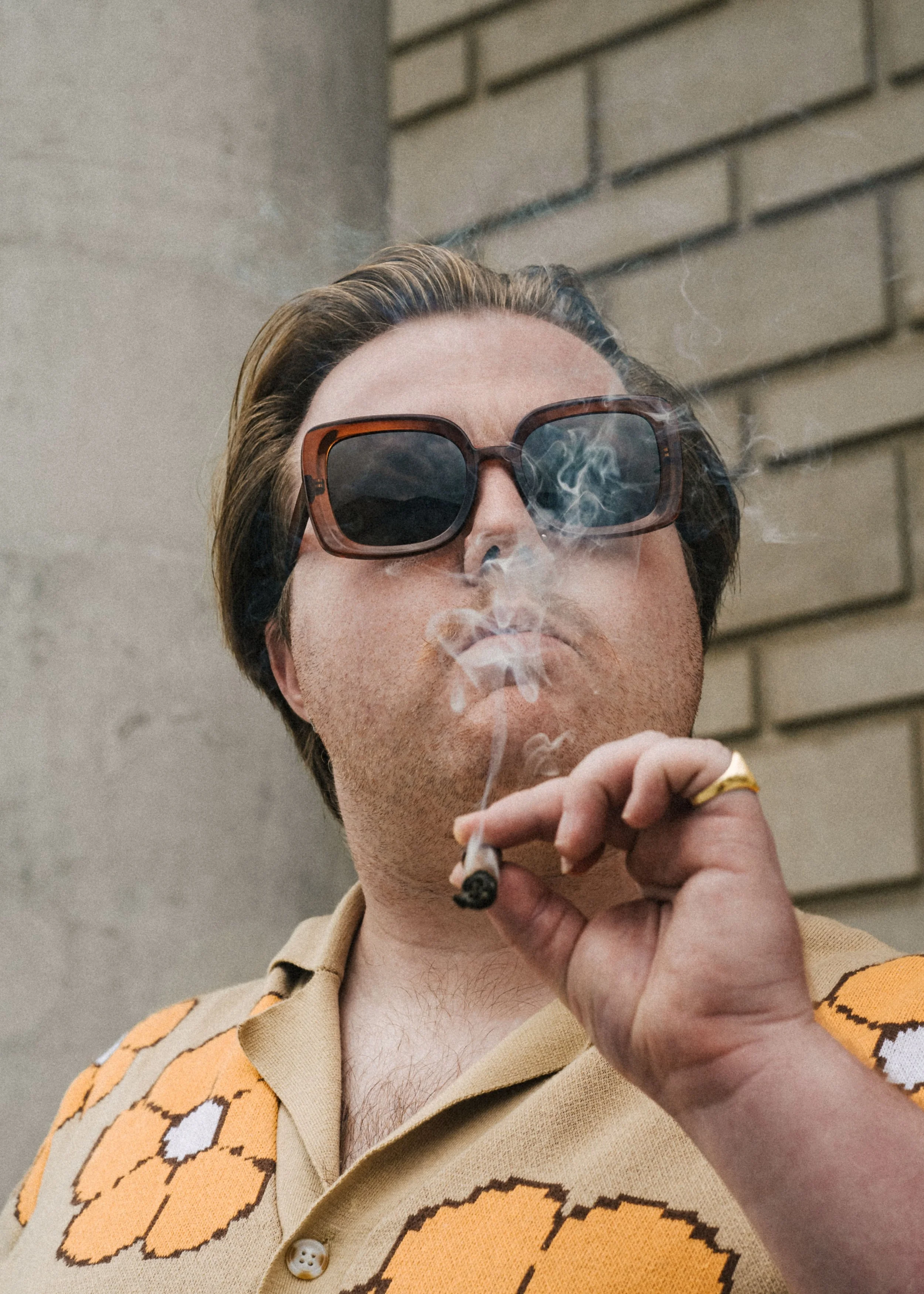 A person with light skin and dark hair wearing large tortoiseshell sunglasses and a beige shirt with an orange and black tiger stripe pattern, smoking a cigarette against a brick wall background.
