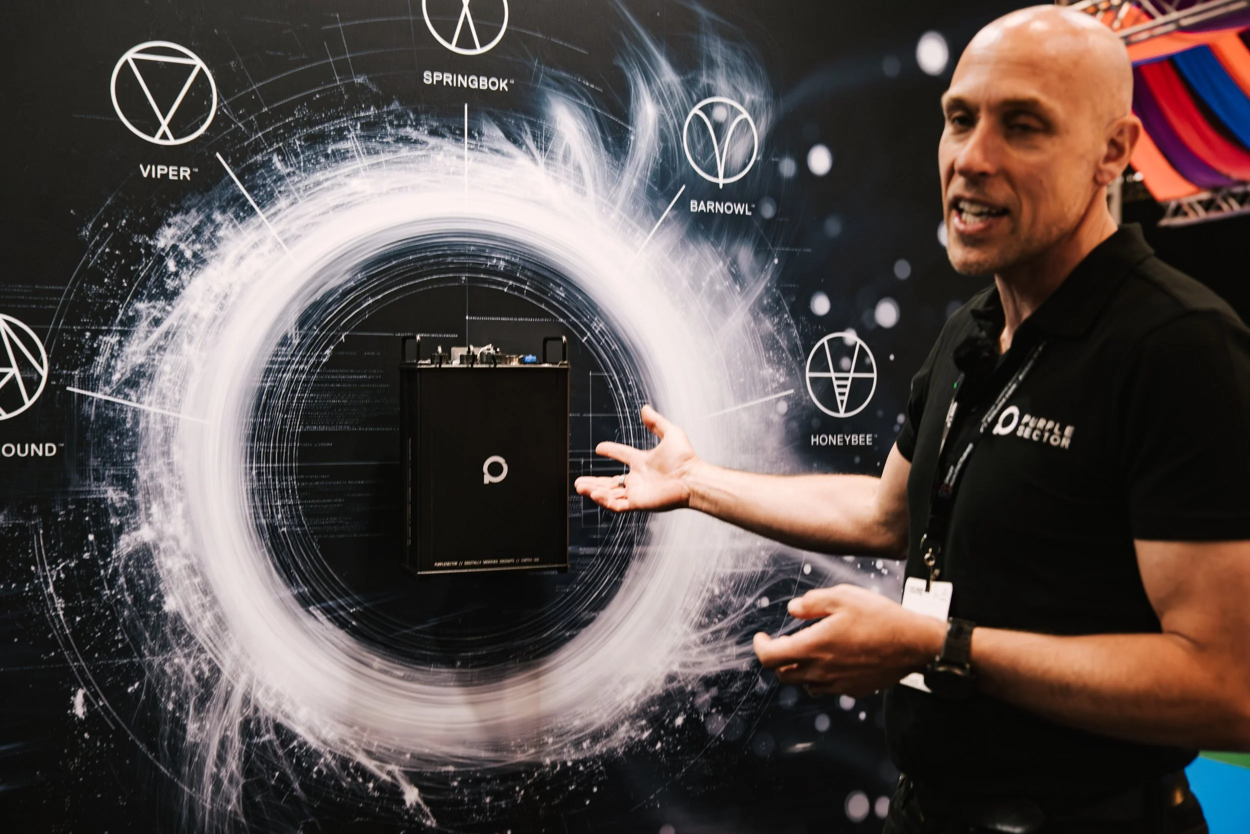 A man is explaining a product at an exhibition, standing next to a black display with a digital illustration of a white, swirling, circular design in the background. The person is wearing a black shirt with some white text, and there are other colorf