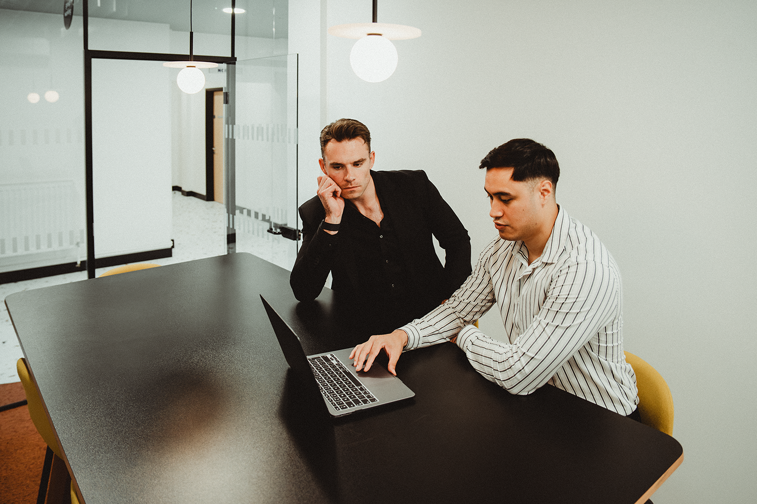 Two men sitting at a black conference table looking at a laptop; one is wearing a striped shirt and the other a black suit jacket, in a modern office space with glass walls and hanging lights.