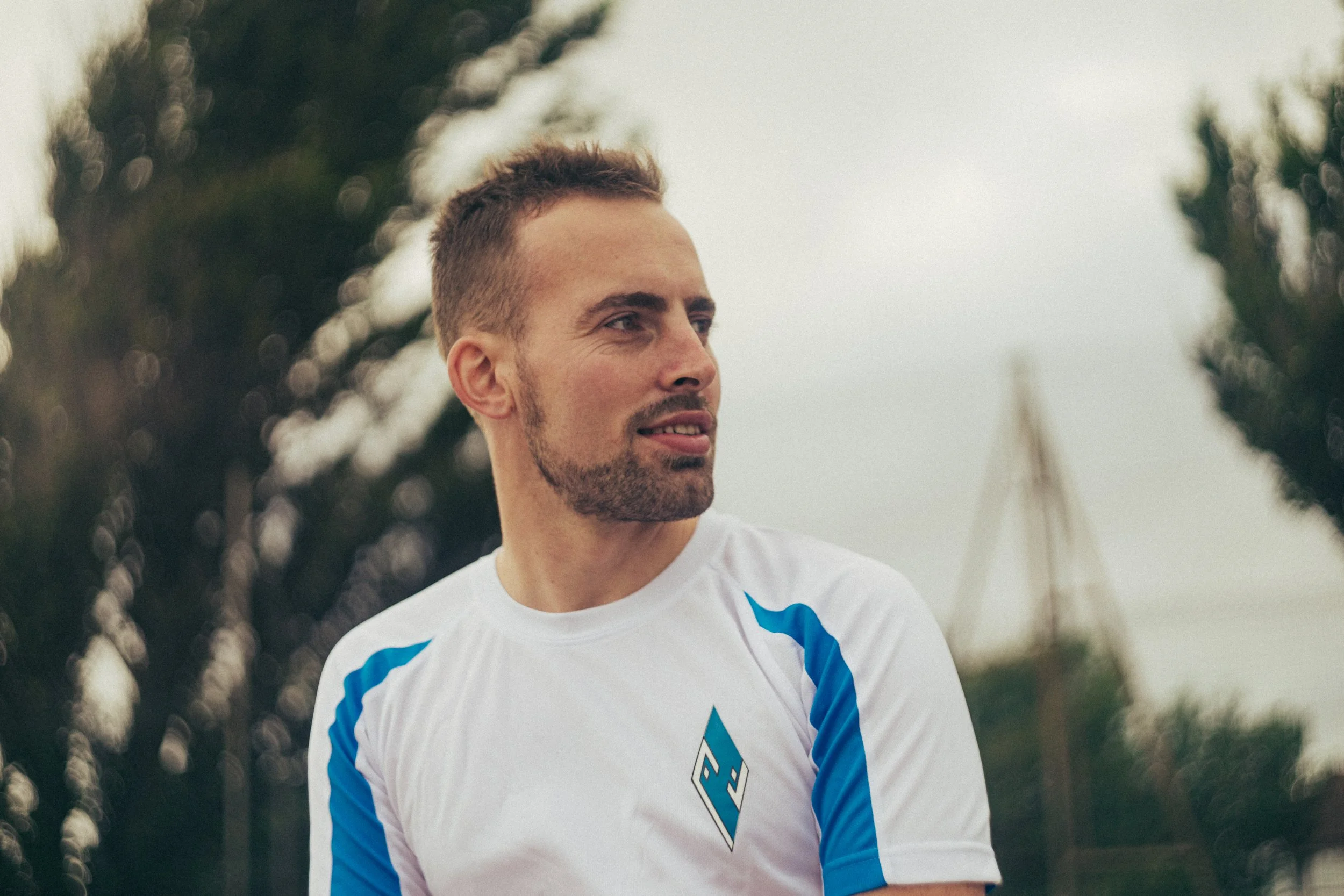 A man with short brown hair, facial hair, and light skin, wearing a white sports jersey with blue accents, standing outdoors with trees in the background.