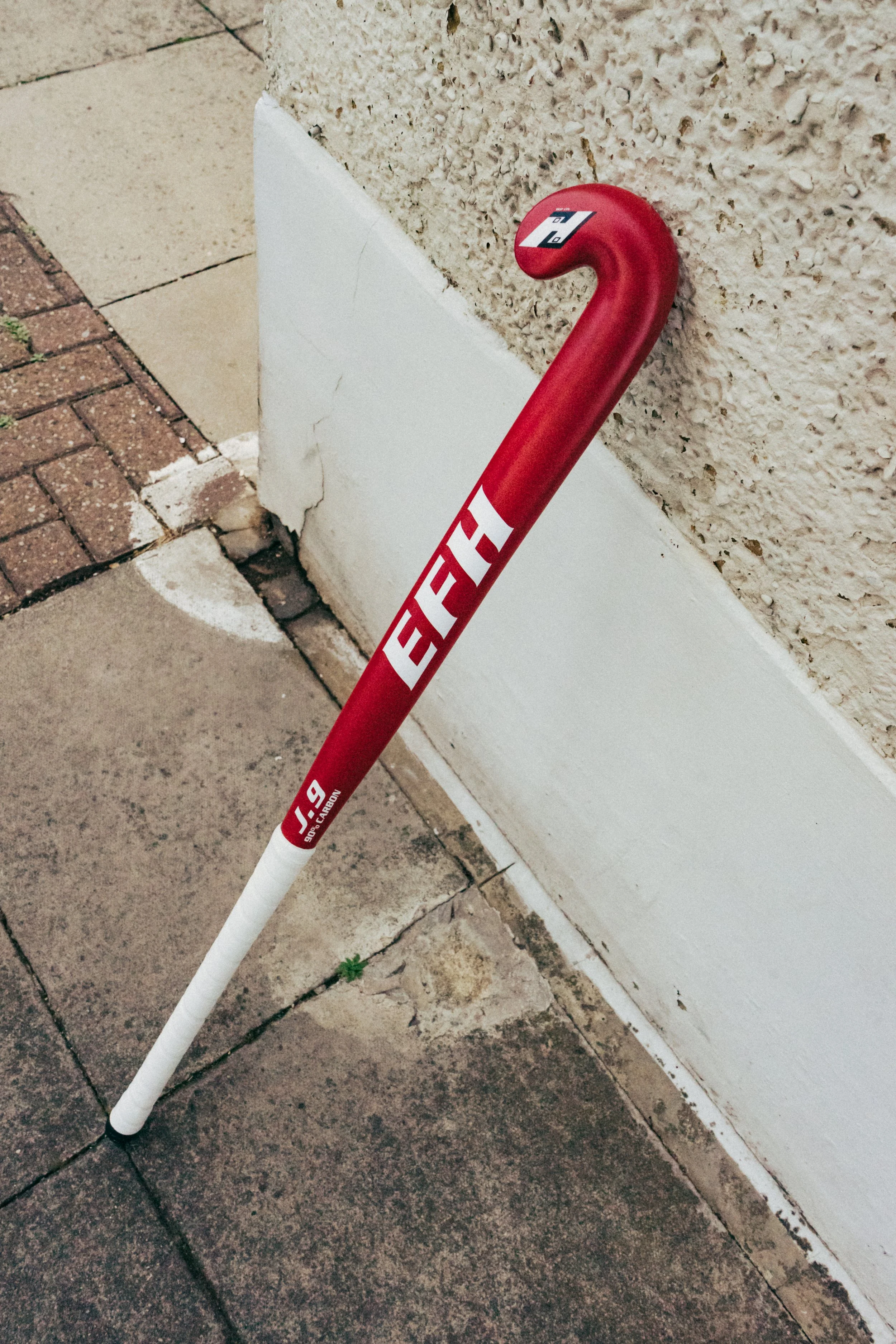 A red and white hockey stick with branding resting against a textured beige wall on a sidewalk.