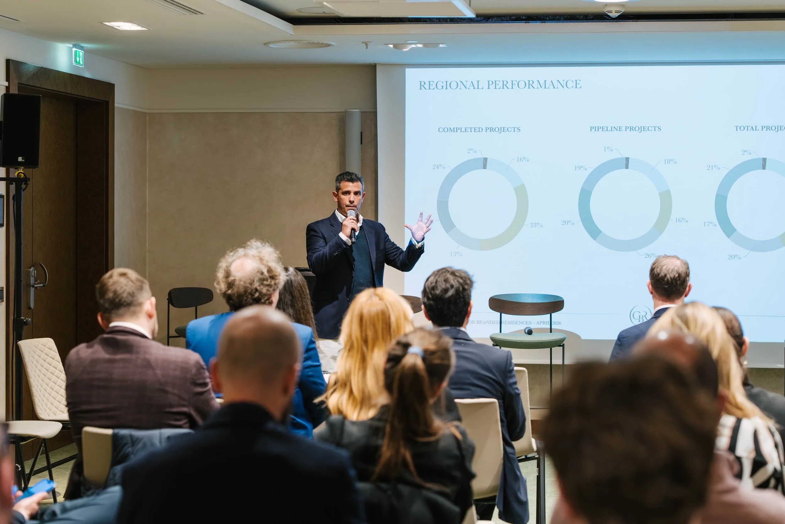 A man in a suit giving a presentation to an audience in a conference room, with a large screen displaying a slide titled 'Regional Performance' and containing pie charts showing project statistics.