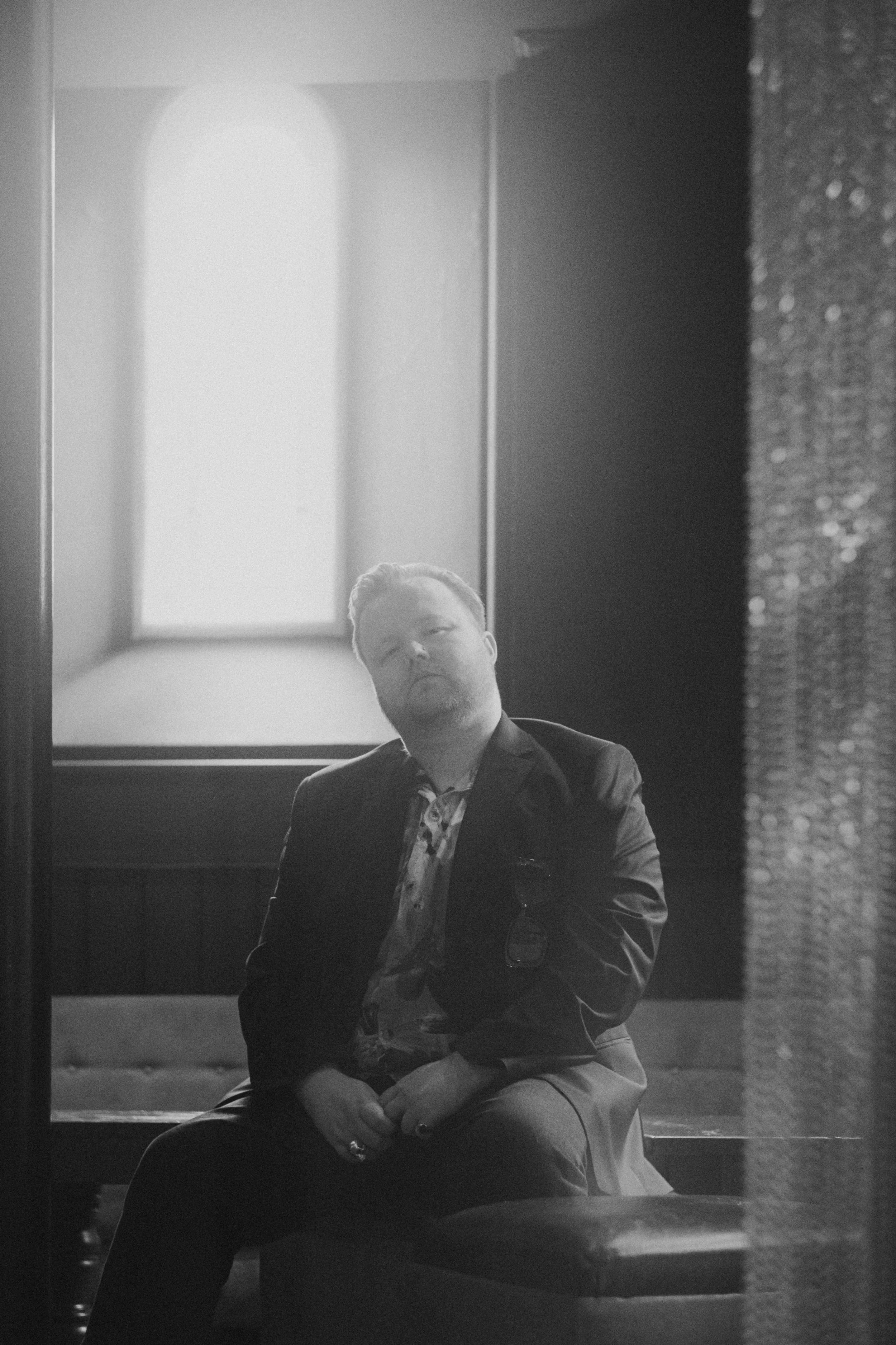 A black and white photo of a man sitting on a bench indoors, with a large window behind him allowing natural light to illuminate the space.