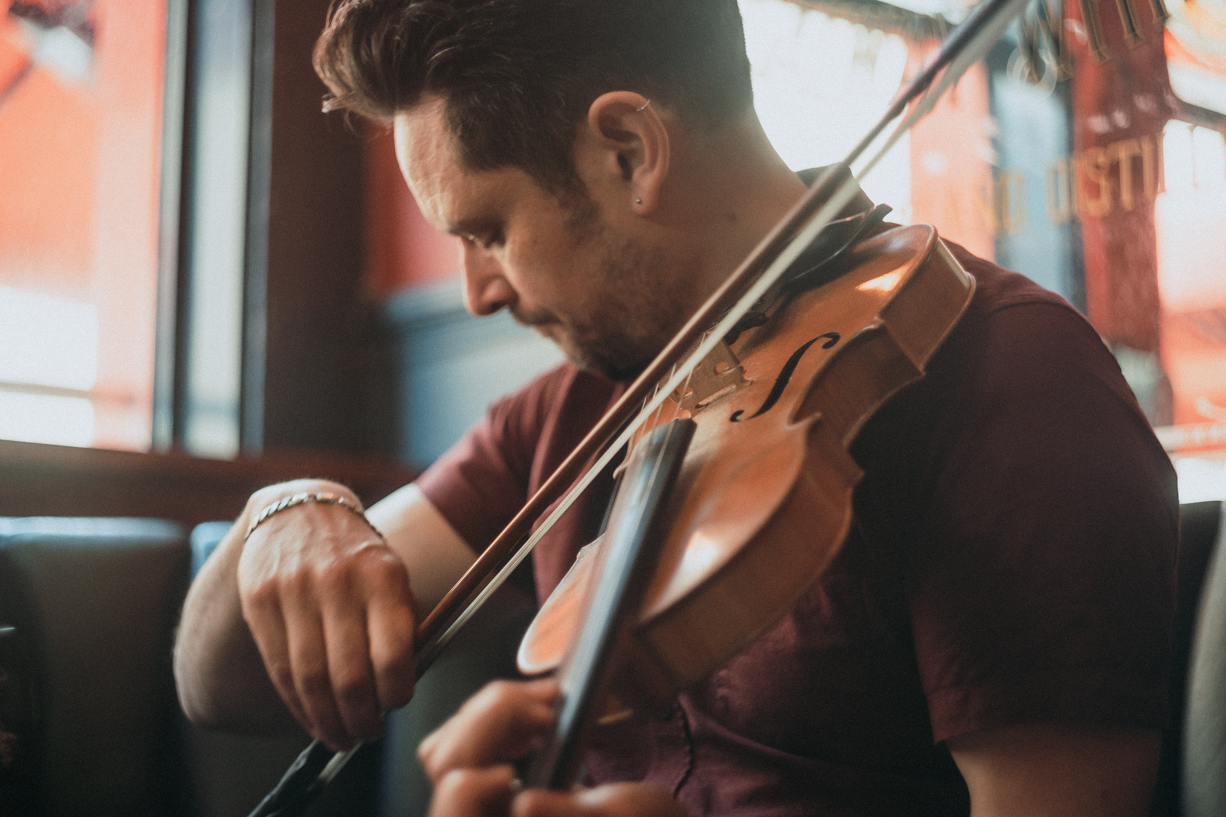 A man with dark hair and earrings playing the violin indoors during daytime, with a red wall and windows in the background.