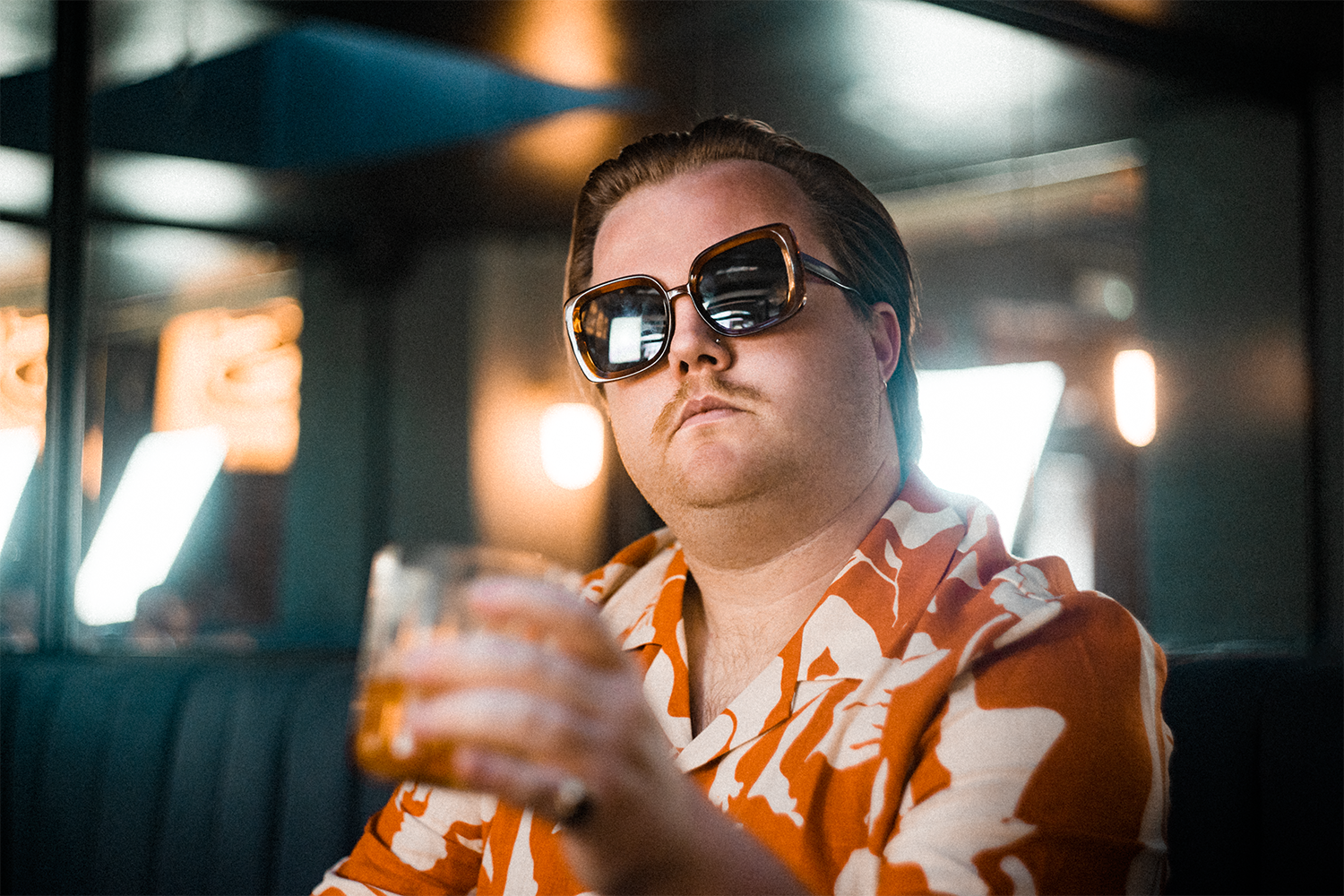 A man wearing large sunglasses and an orange and white patterned shirt, holding a glass of amber-colored beverage in a dimly-lit bar or restaurant